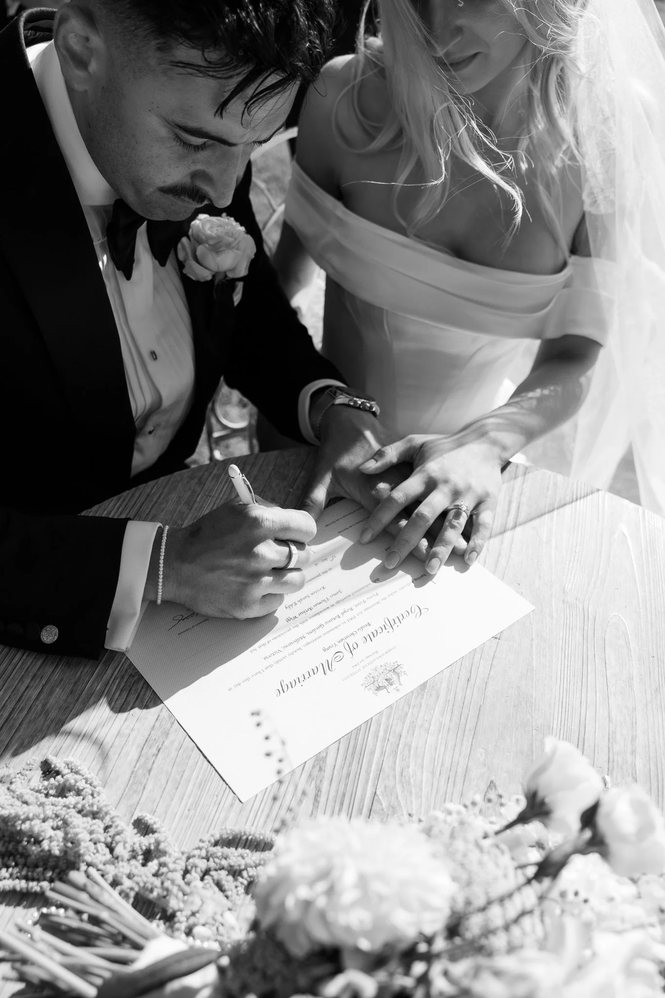 A black and white photo of a man signing a wedding certificate with a woman standing beside him, holding his hand, at a wedding ceremony.