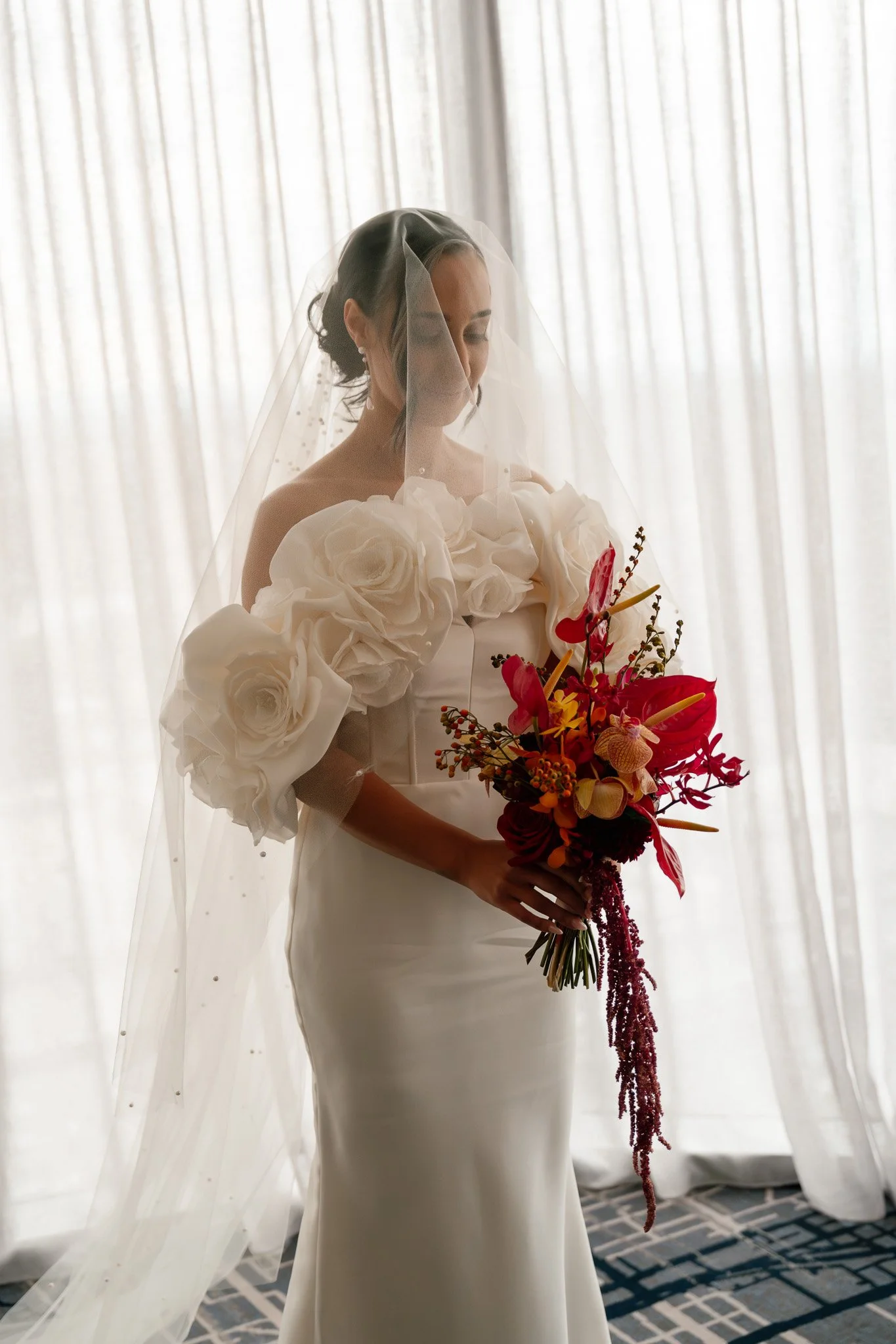 A bride in a white wedding gown with large floral sleeves, holding a bouquet of red and orange flowers, standing in front of sheer curtains.