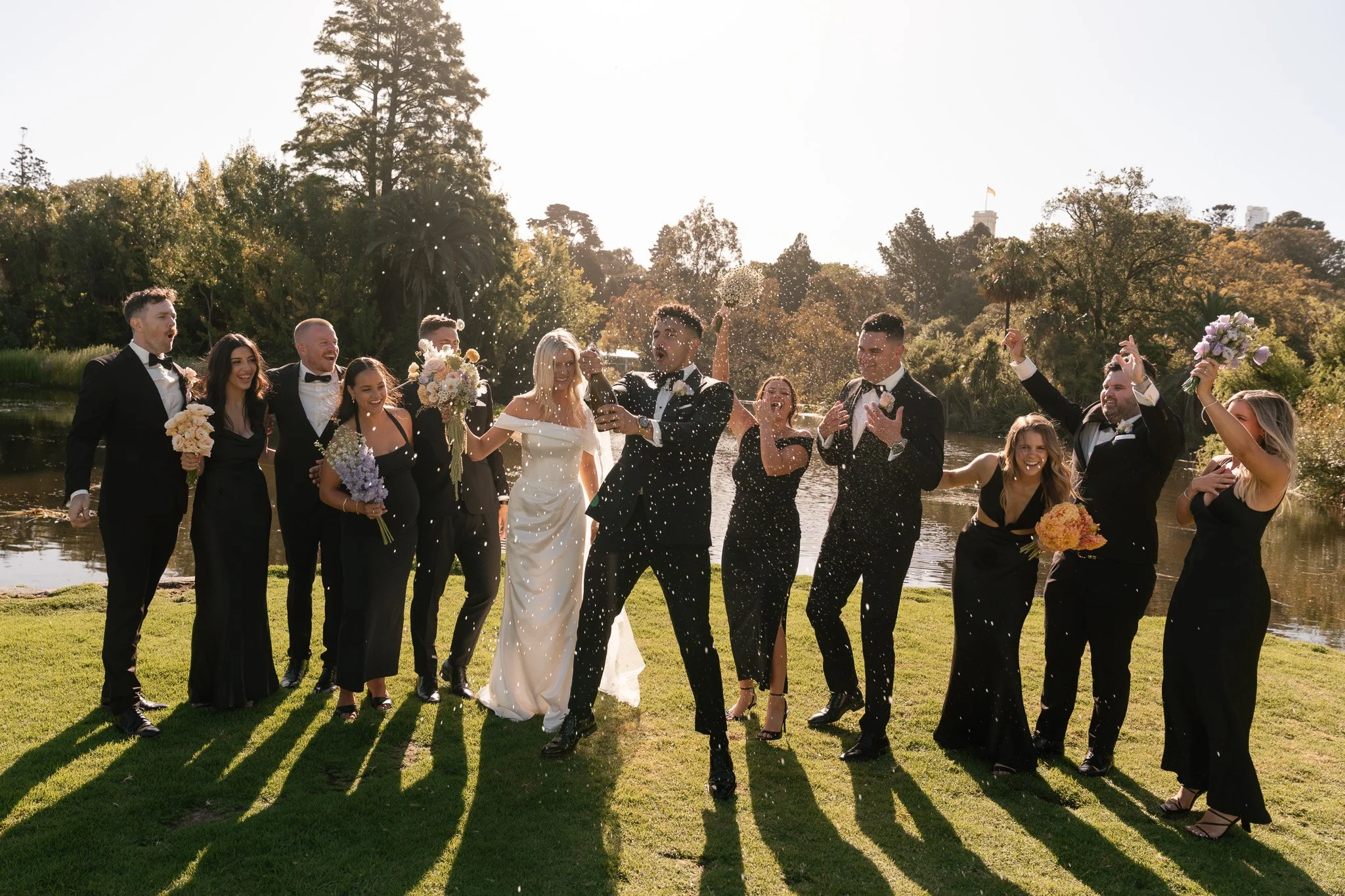 Group of wedding guests celebrating outdoors by a pond, with some throwing confetti, during daytime.