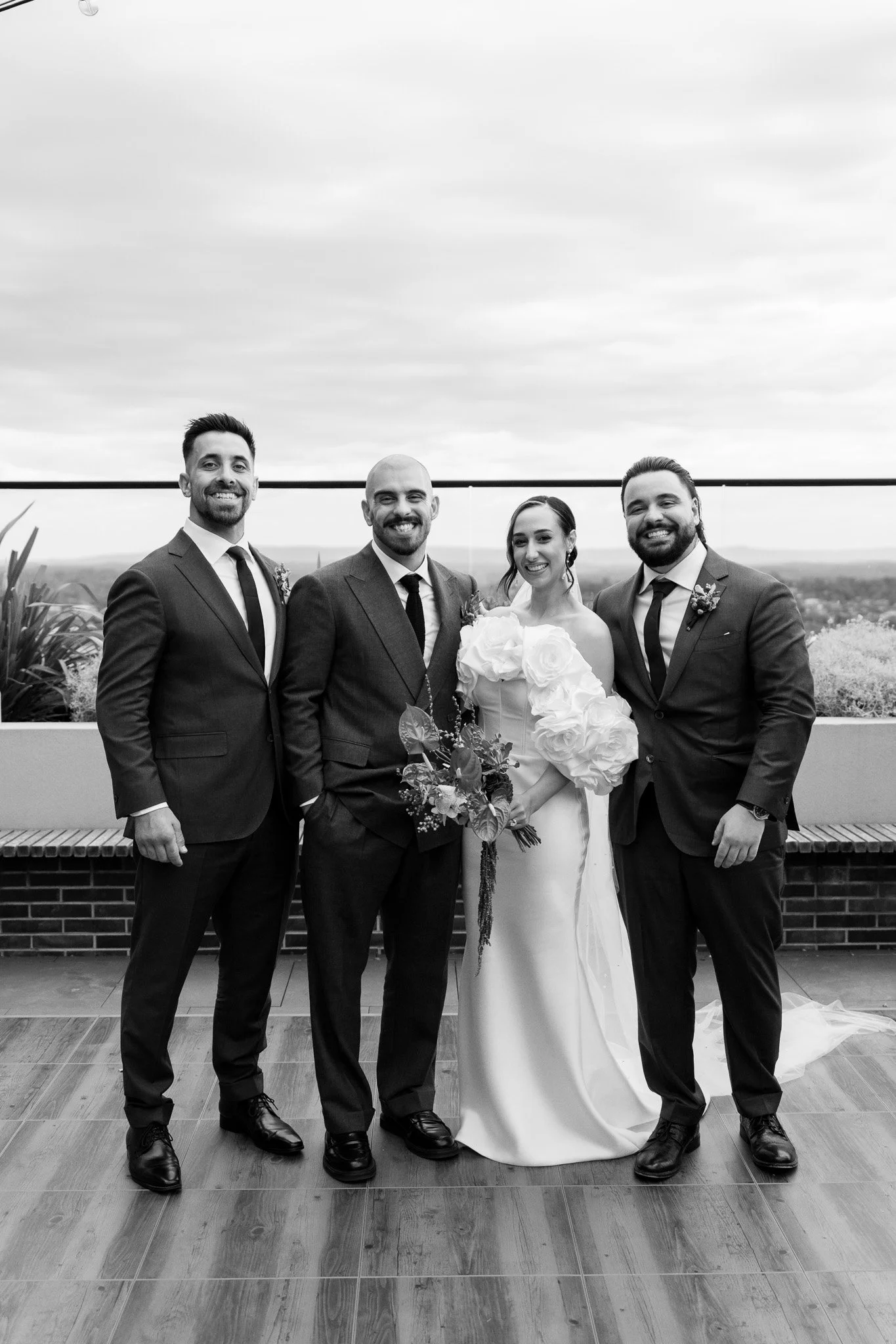 Black and white photo of a wedding party with the bride, groom, and two groomsmen, all dressed in formal attire, standing outdoors on a rooftop with a scenic landscape in the background.