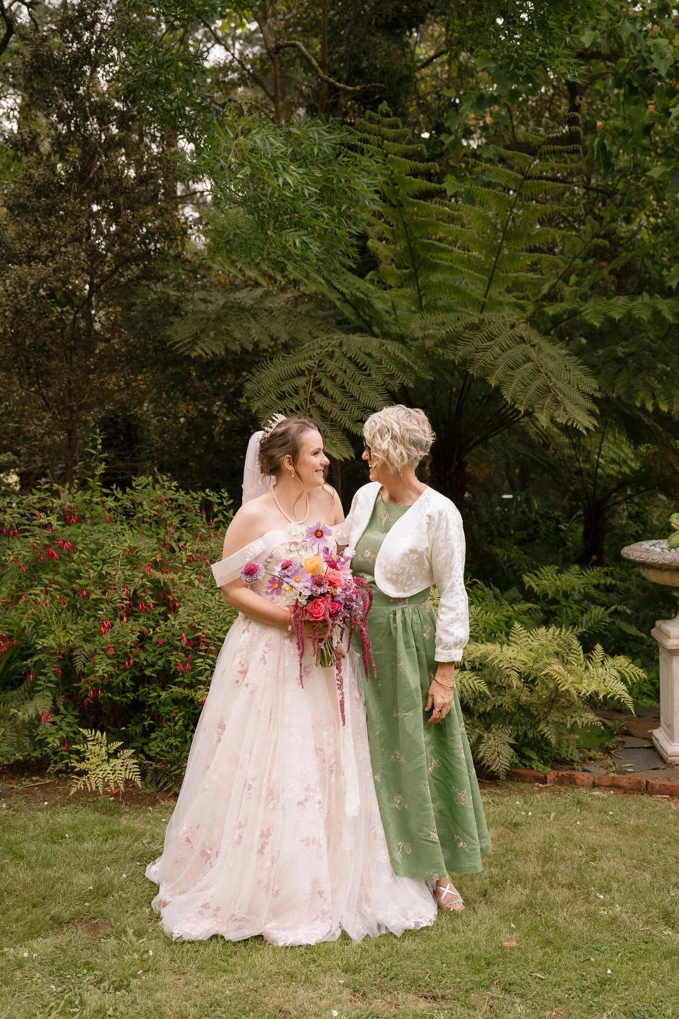A bride in a wedding dress holding a bouquet, standing next to an older woman in a green dress and white jacket, in a lush garden setting.