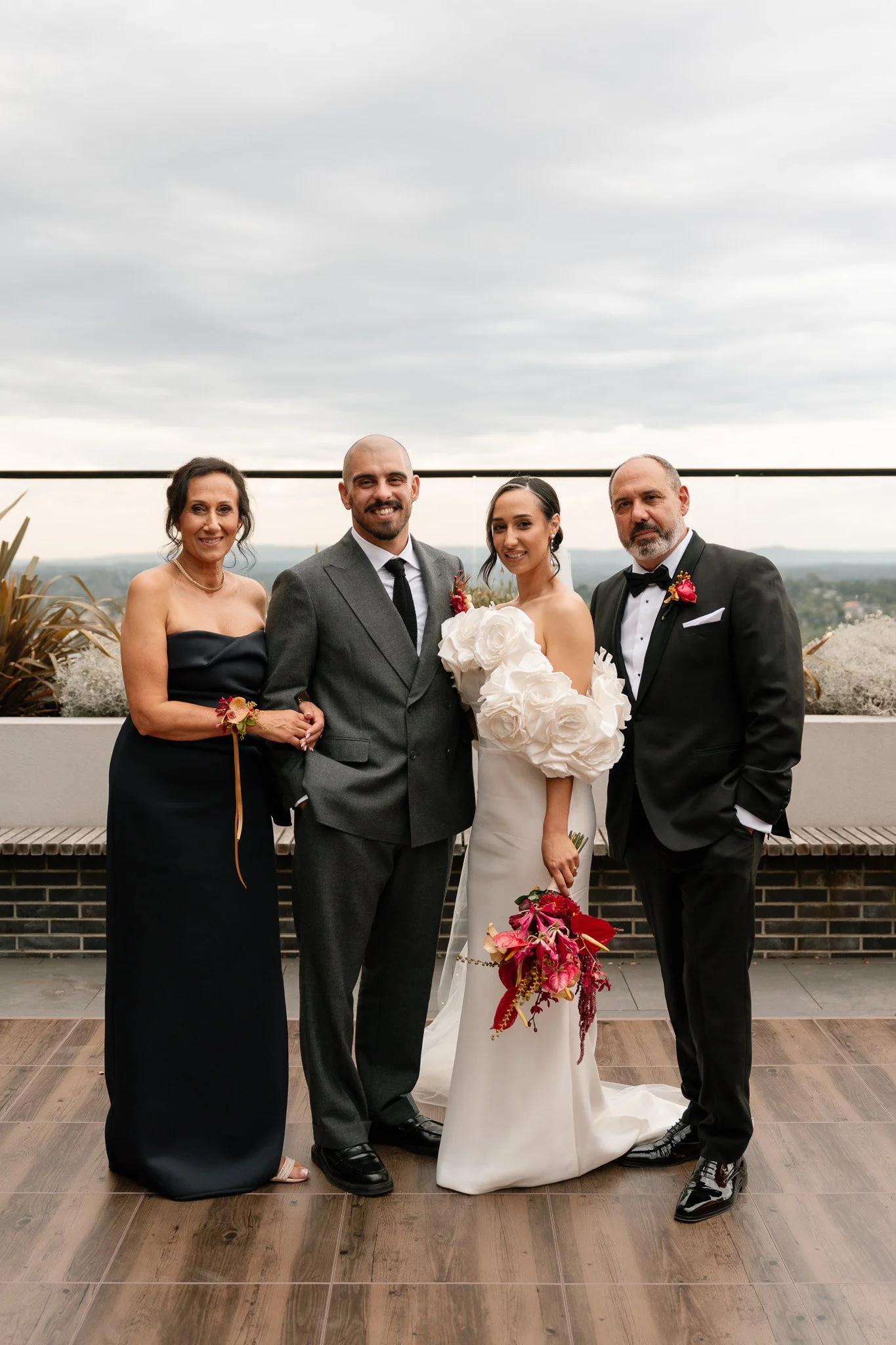 Group of four people, two women and two men, standing outdoor on a balcony during a wedding. The woman in the middle is wearing a white wedding gown with large flower details on her sleeves and holding a colorful bouquet. The man next to her is in a 