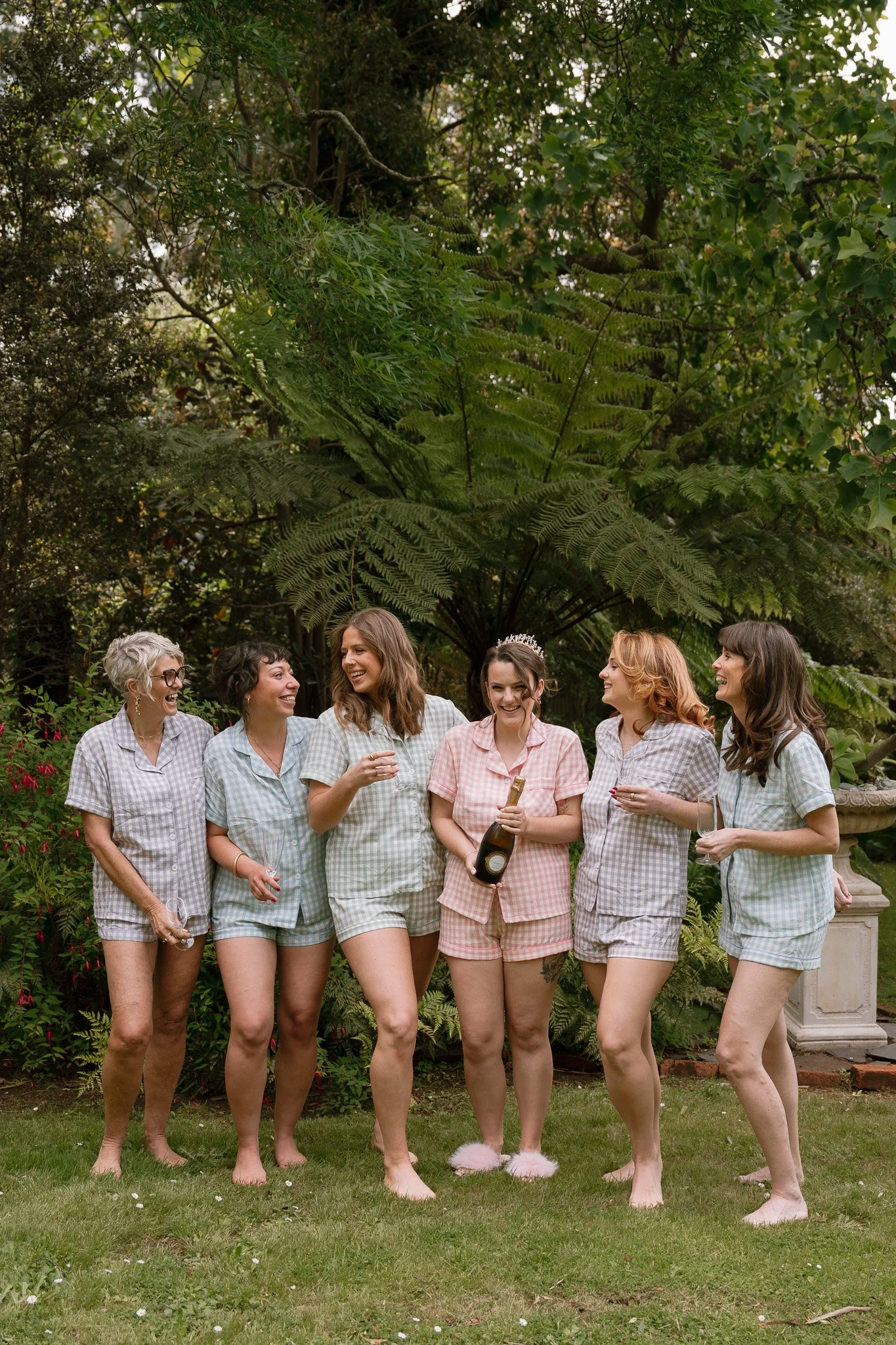 Group of women celebrating in pajama shirts outdoors, one holding a bottle of champagne, others holding glasses, smiling and chatting, in a lush garden setting.