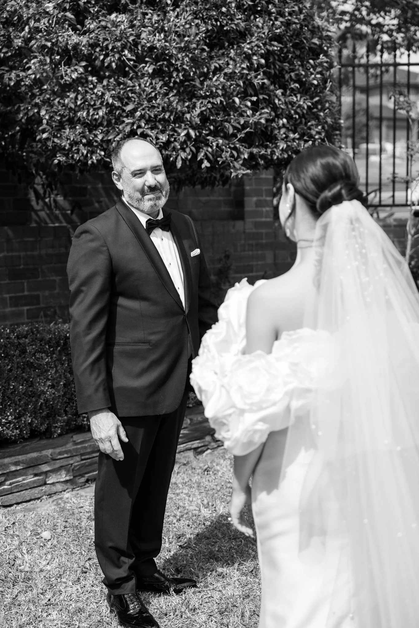 A man in a tuxedo faces a woman in a wedding dress outdoors near a brick wall and a tree.