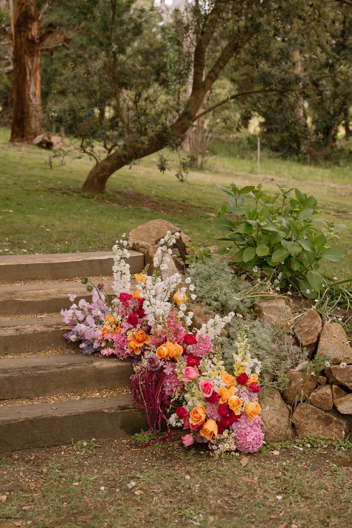 Colorful flower arrangement on outdoor wooden steps in a lush garden with trees and greenery in the background.