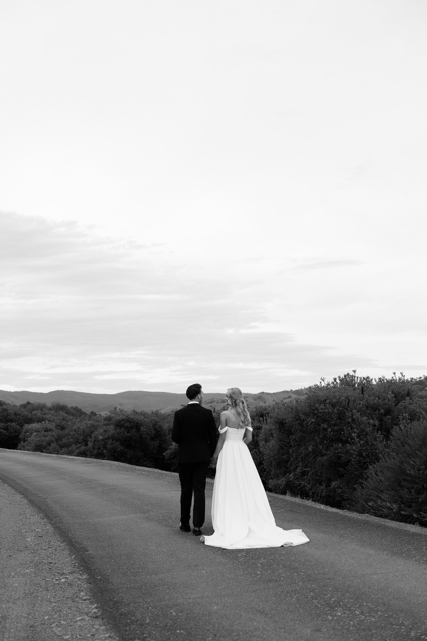 Black and white photograph of a bride and groom holding hands, walking on a rural road with hills and trees in the background.