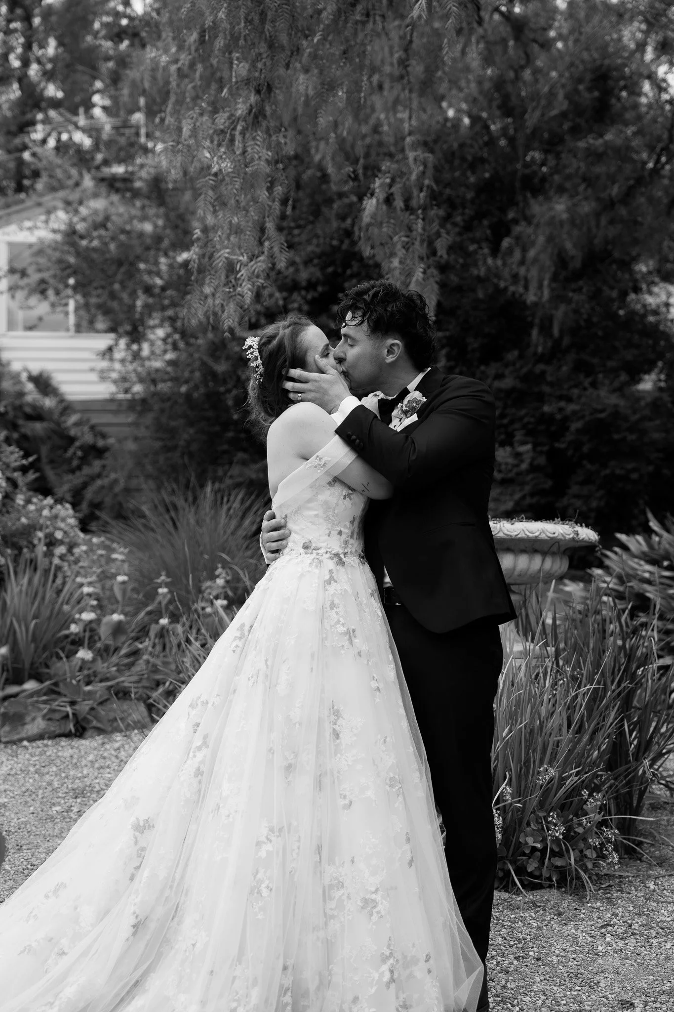 A black and white photo of a newlywed couple sharing a tender kiss outdoors, with lush foliage in the background.