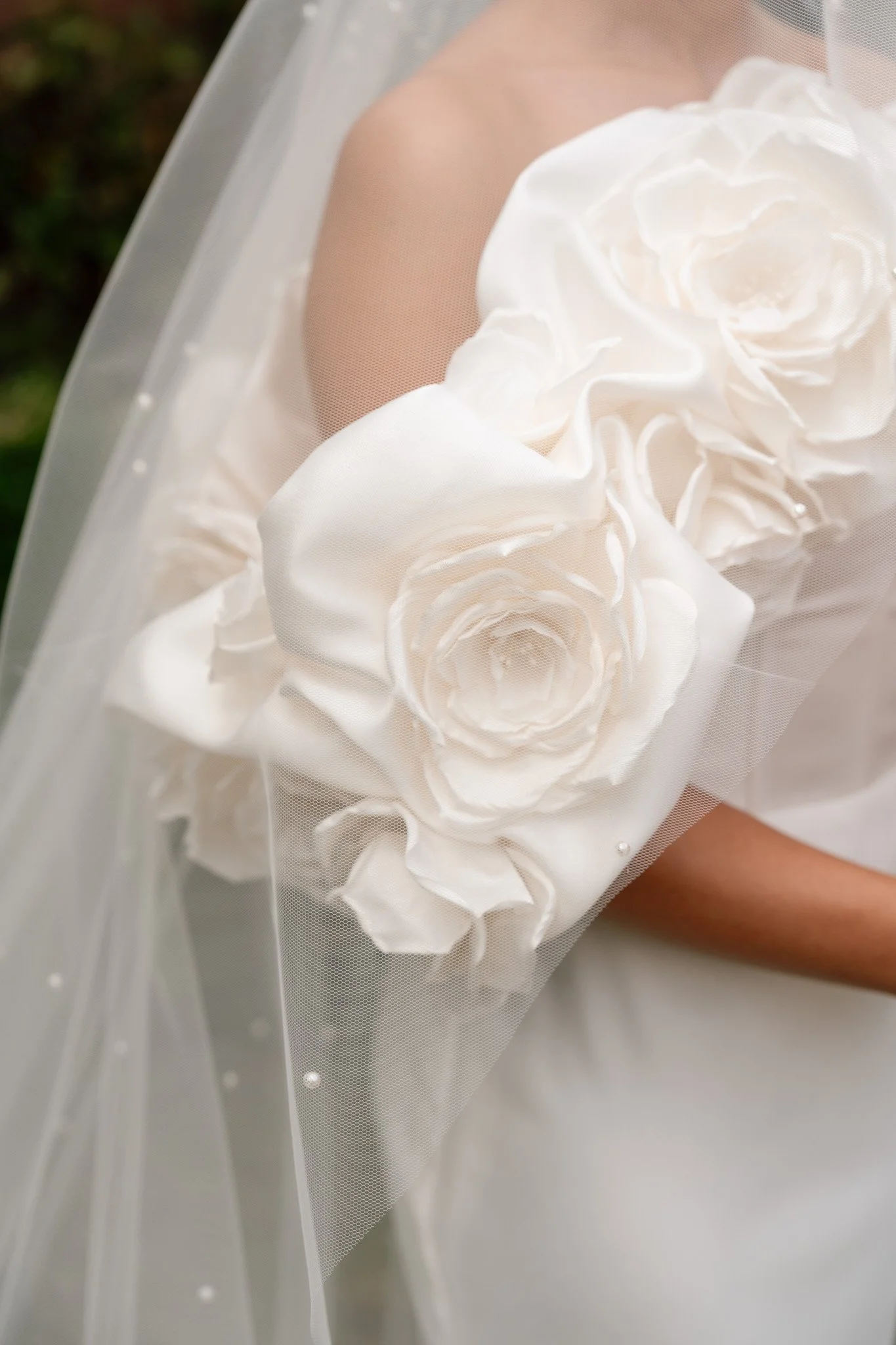 Close-up of a woman's arm and shoulder with a white dress adorned with large fabric roses, and a sheer veil with small pearls.