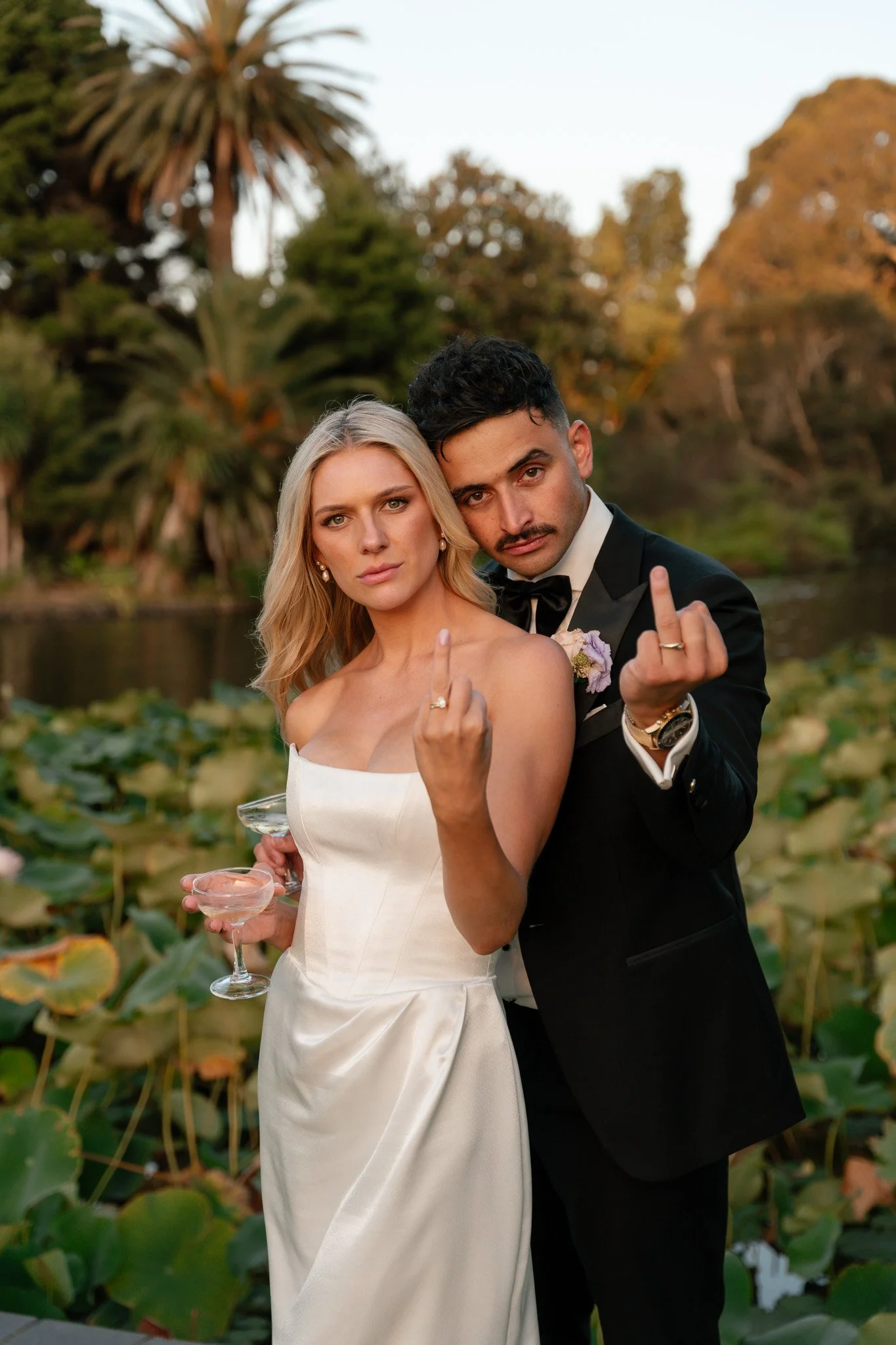 A wedding couple making rude hand gestures while standing outdoors near water lilies.