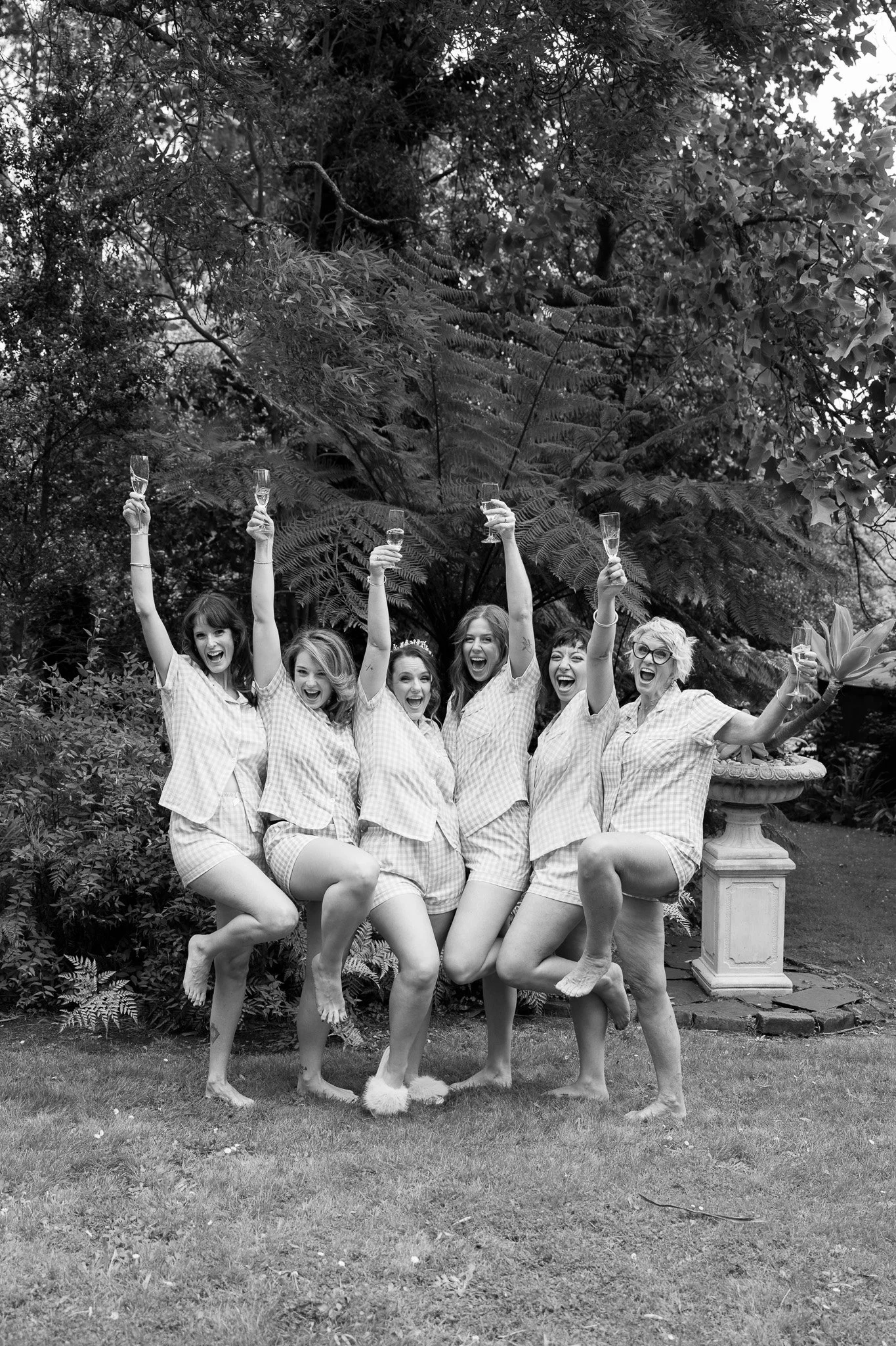 A group of six women in pajamas celebrating, holding champagne glasses, and lifting their legs in a garden or backyard with trees and plants in the background.