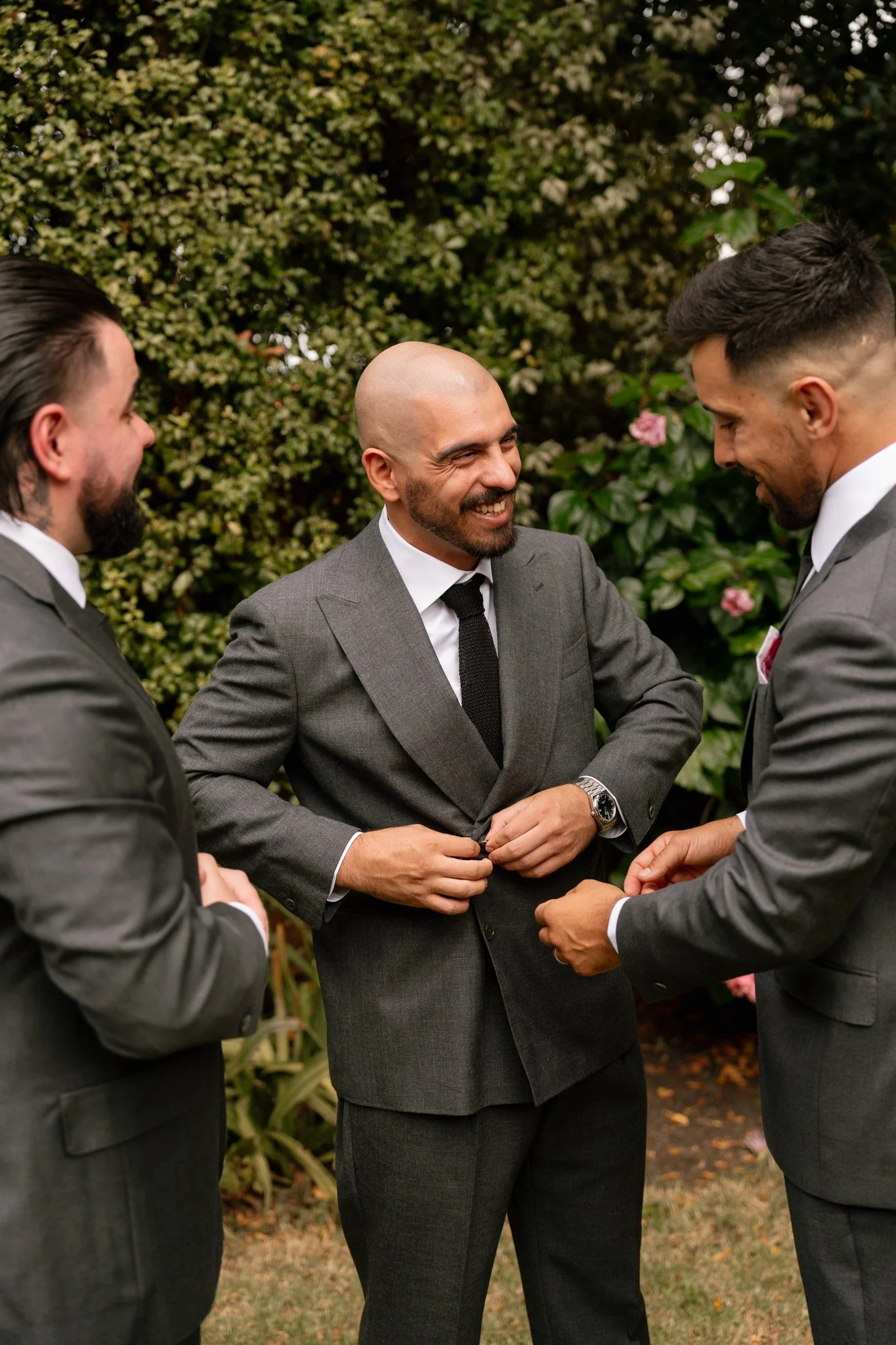 Three men dressed in suits, two with dark hair and one with a shaved head, standing outdoors and smiling at each other.