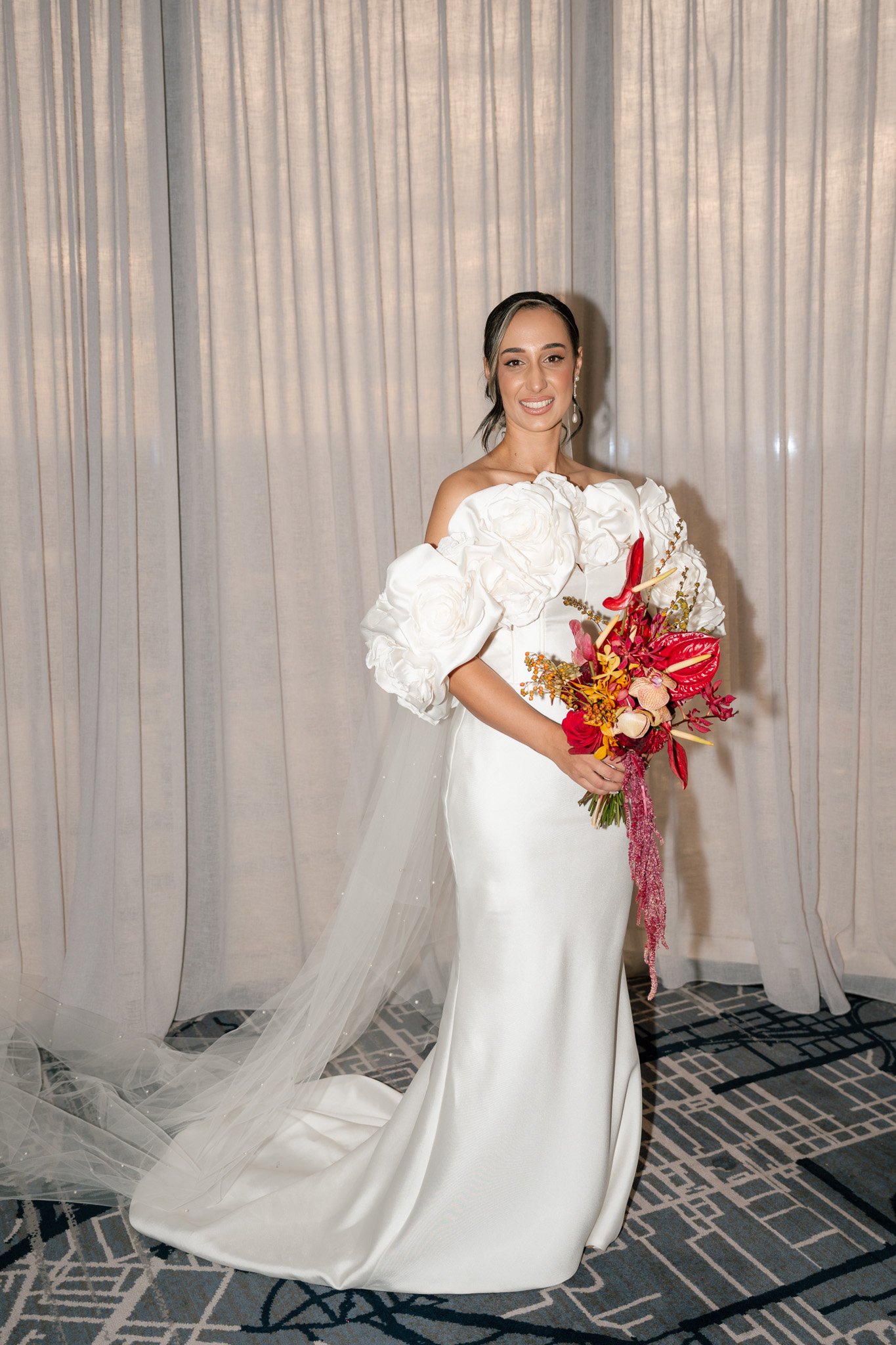 Bride in white wedding dress holding a bouquet of red, pink, and orange flowers, standing in front of light-colored curtains.