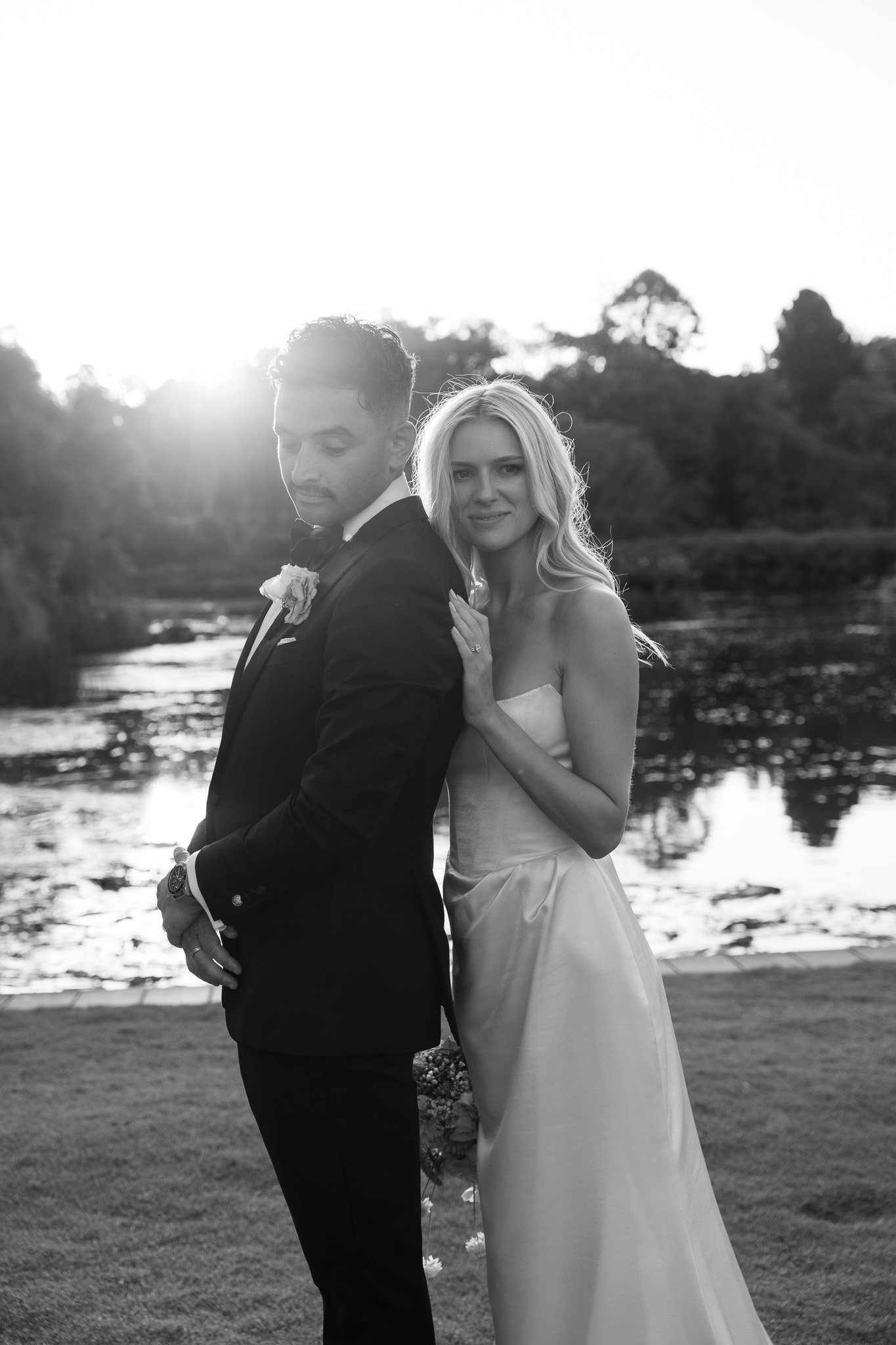 Black and white photo of a bride and groom by a river, with the sun setting behind trees, the bride touching the groom's shoulder.