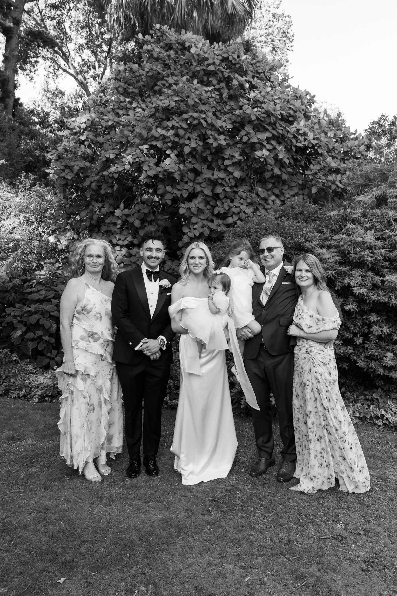 Black-and-white photo of seven people outdoors, including a bride in a white gown holding a young girl in a wedding dress, a groom in a tuxedo, and four adults, standing on grass with dense trees in the background.