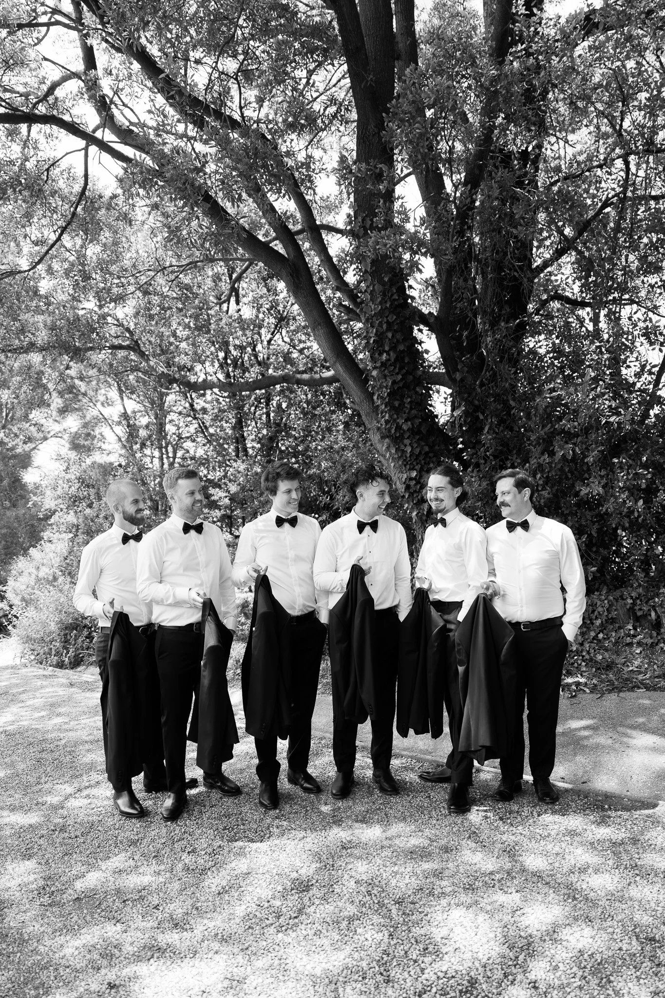 Six men dressed in formal shirts and bow ties holding their tuxedo jackets, standing outdoors beneath a large tree, smiling and engaging in conversation.