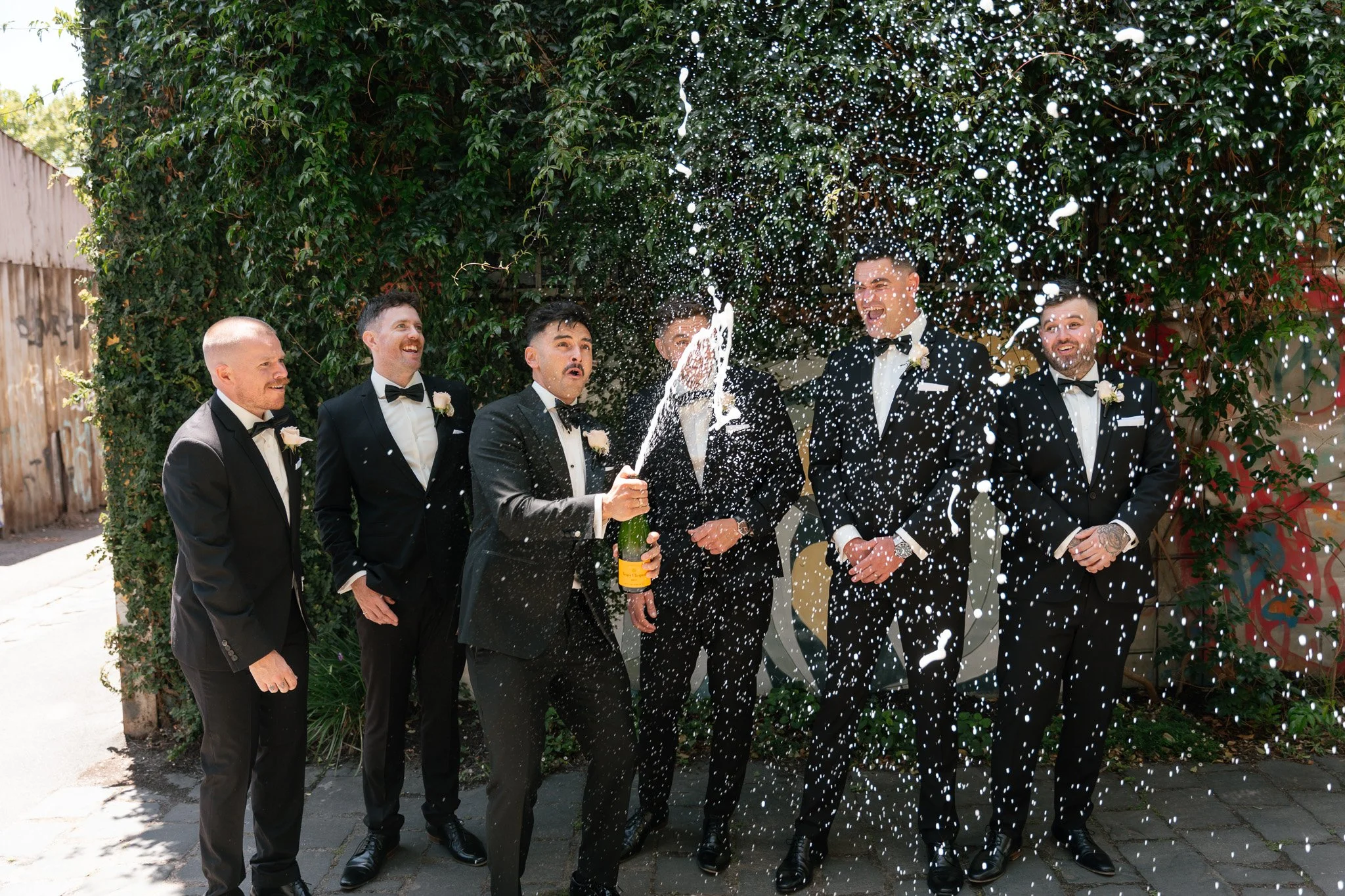 Groom and groomsmen celebrate with champagne and confetti outdoor on a sunny day.