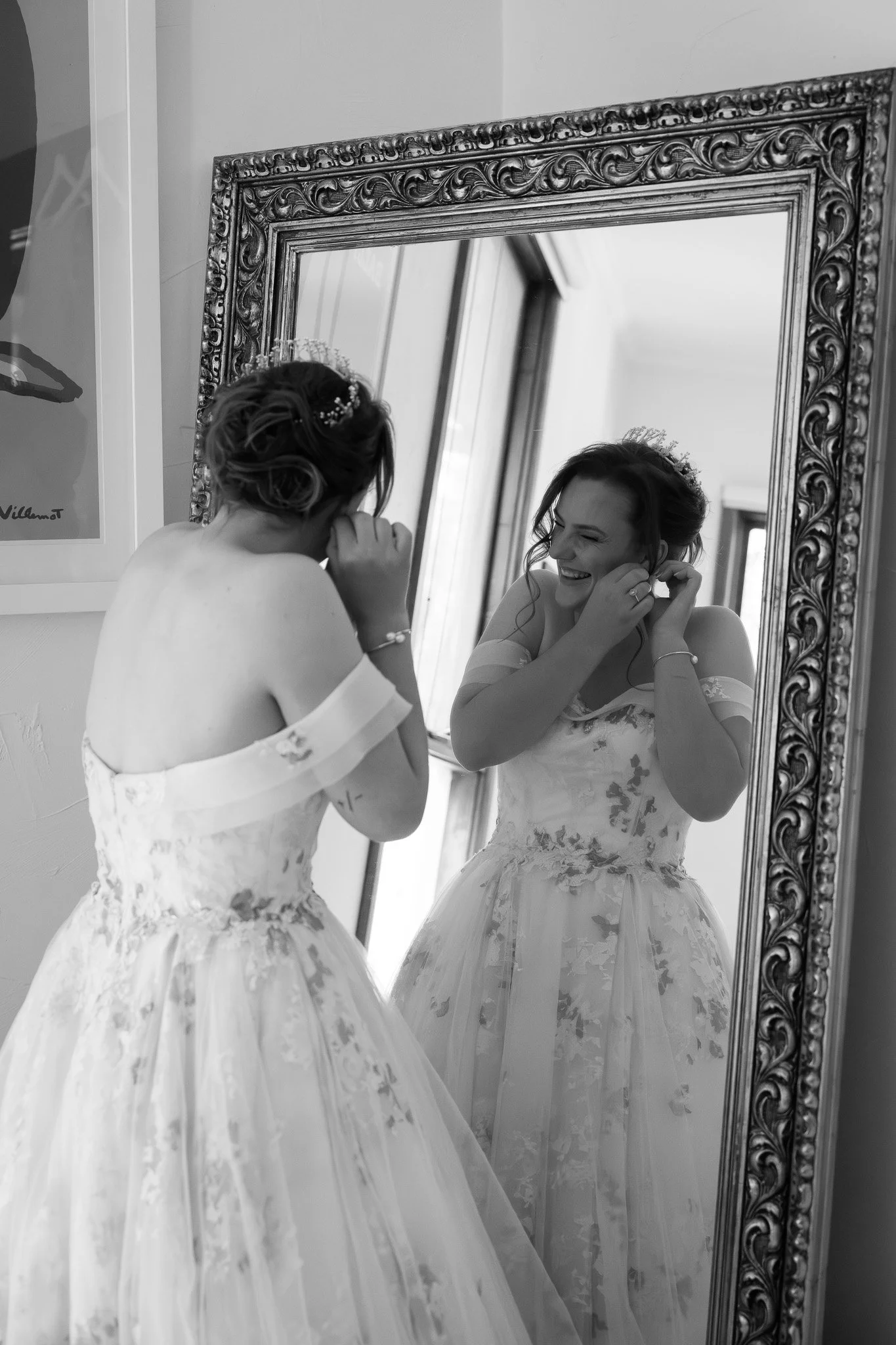 A woman in a wedding dress is smiling and adjusting her earring in front of a large ornate mirror.