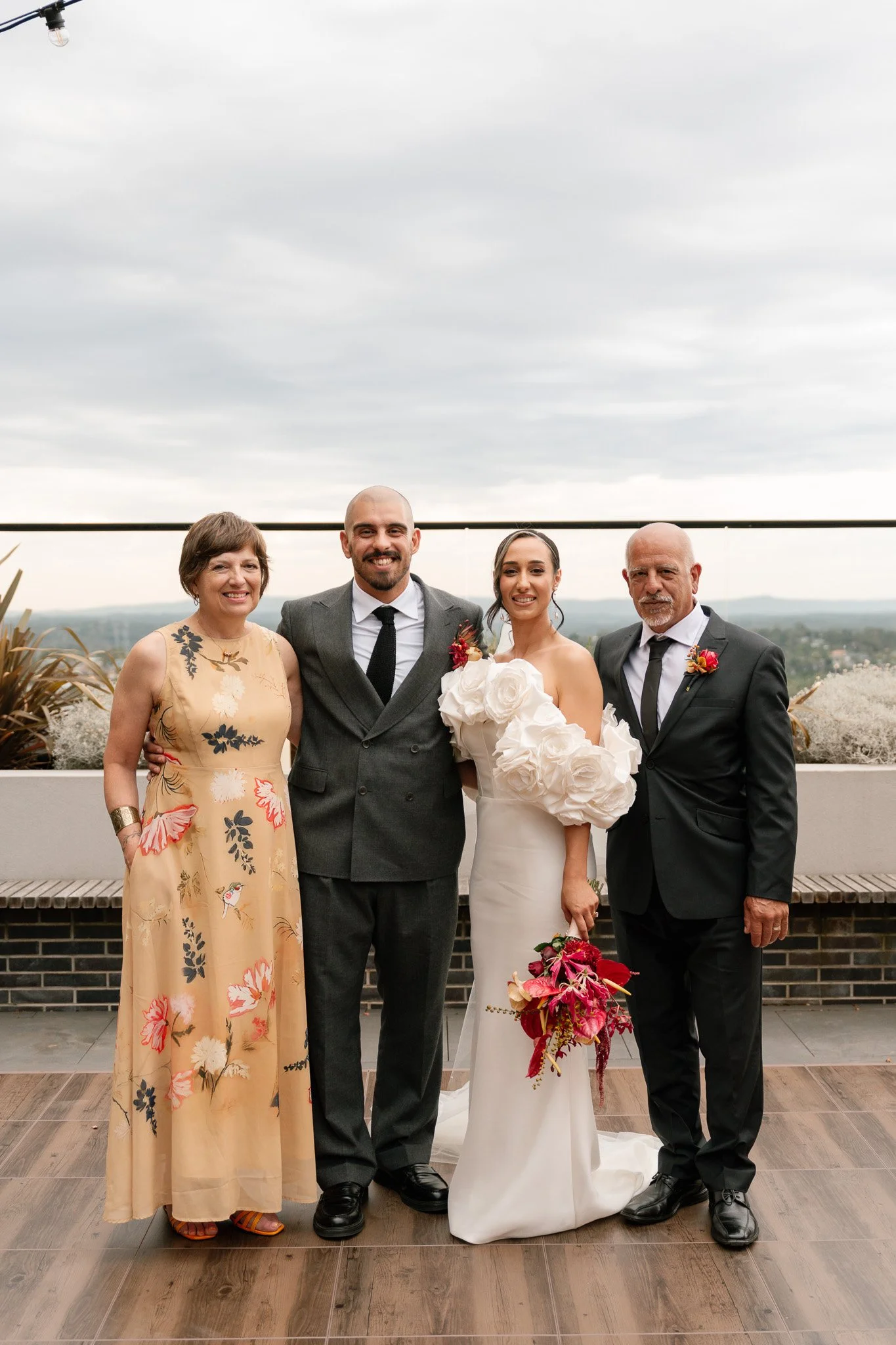 Group of four people at a wedding, including a bride in a white gown holding a bouquet, and three others in formal attire, standing outside with a city view in the background.