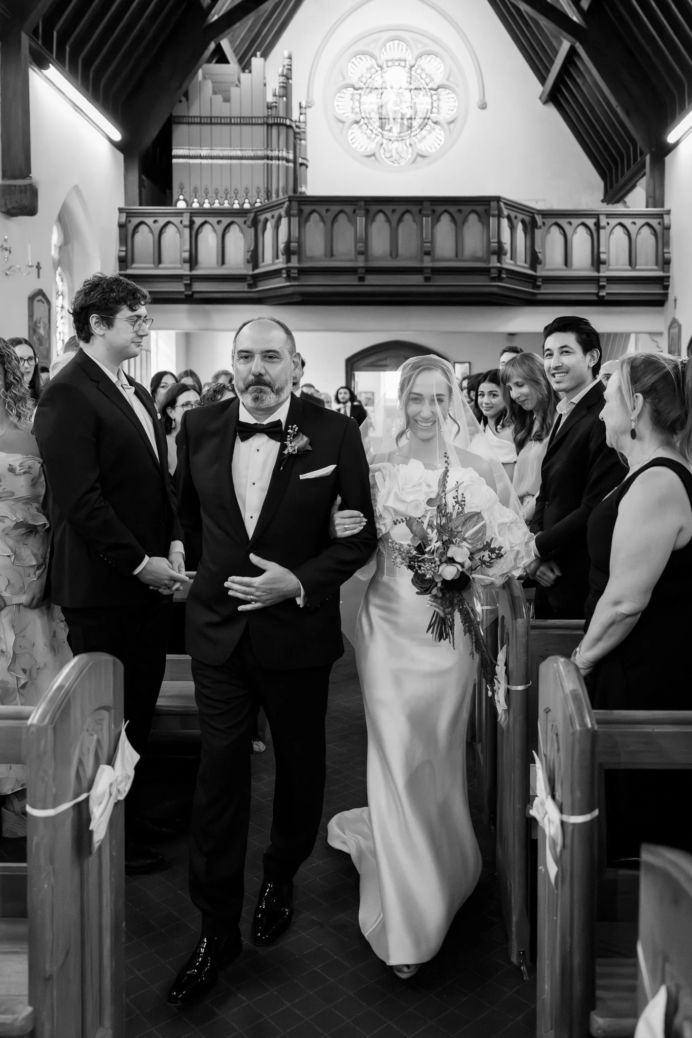 A bride walking down the aisle in a church, accompanied by a man, possibly her father, with guests seated on both sides smiling and watching during a wedding ceremony.