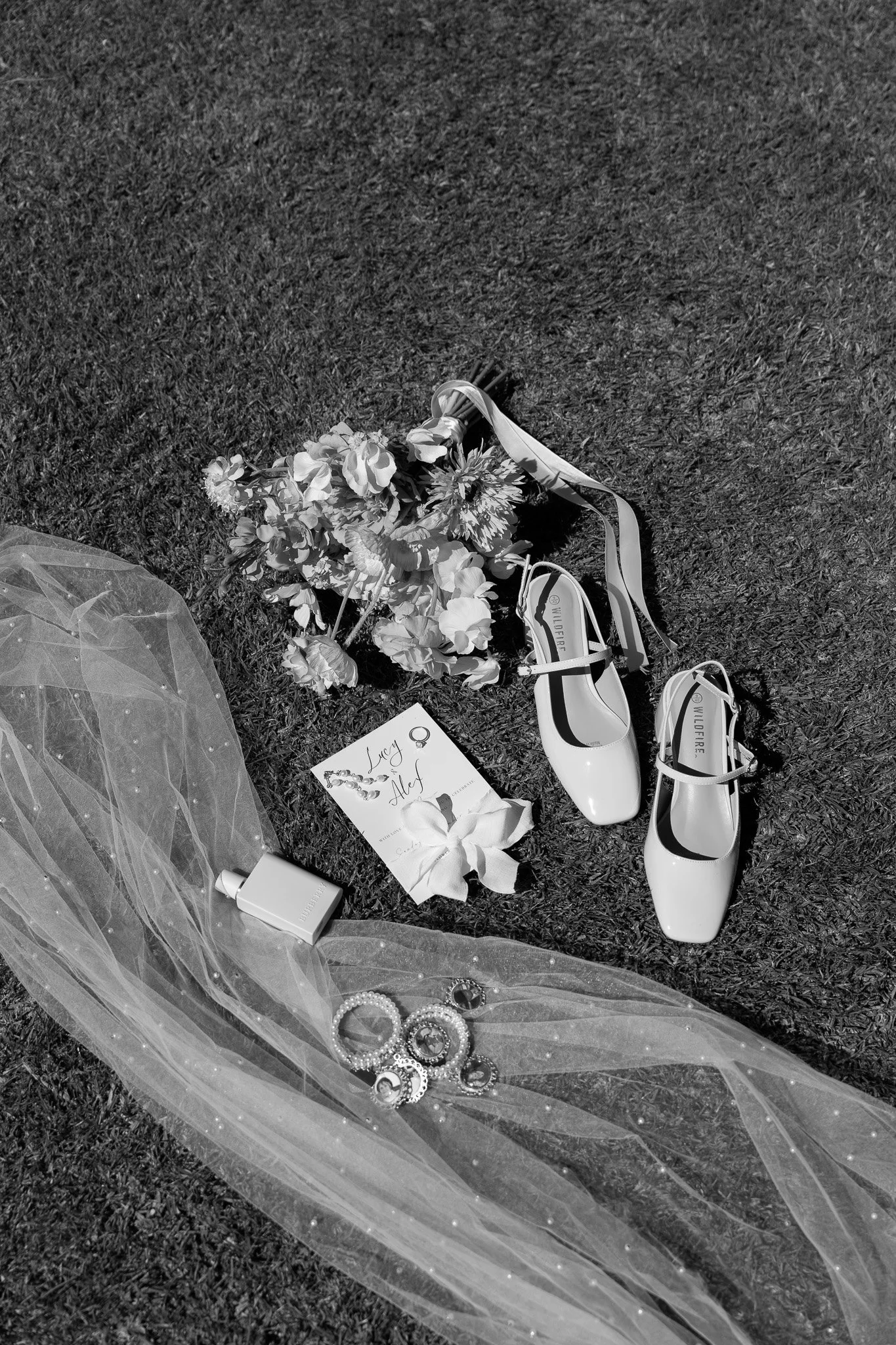 Wedding accessories including a bouquet, a pair of white high heels, a veil, a perfume bottle, jewelry, and a handwritten note on grass.