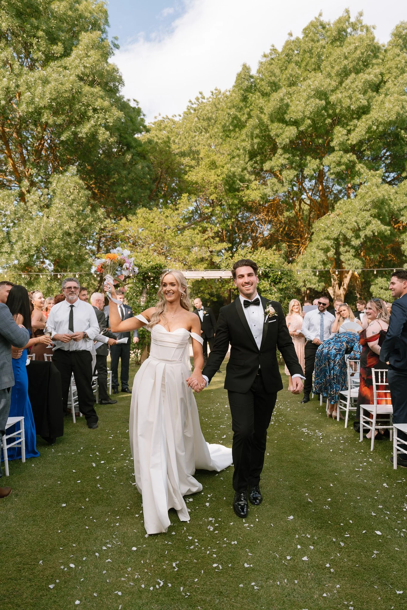 A newly married couple walking hand in hand down the aisle at their outdoor wedding, surrounded by friends and family celebrating.