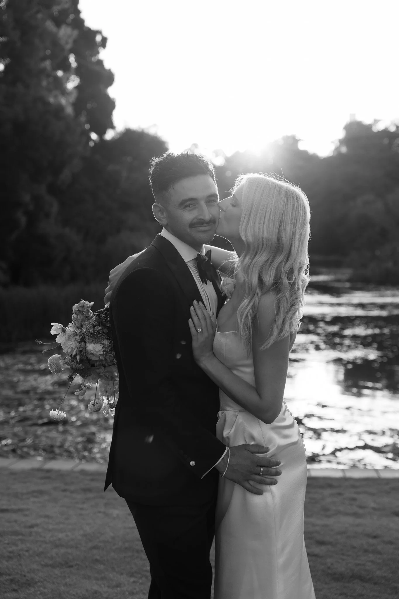 Black and white photo of a bride and groom standing outdoors, with the bride kissing the groom on the cheek. The groom is dressed in a tuxedo holding a bouquet, and the bride is in a satin dress. Behind them, there is a body of water and trees, with 