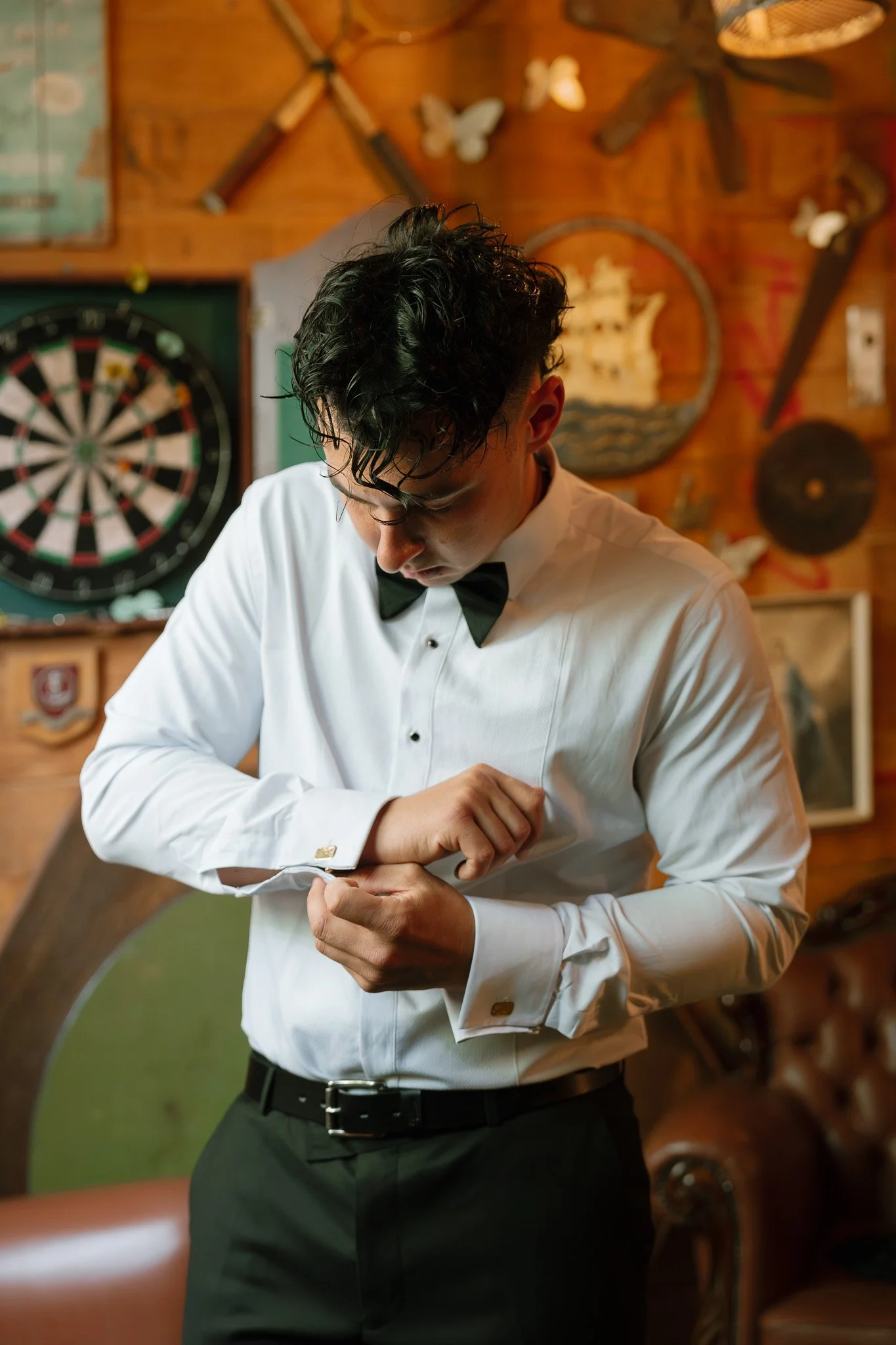 A young man in a white tuxedo shirt with a black bow tie adjusting his cufflink in a room decorated with vintage decor, including a dartboard, butterfly wall decorations, and a leather sofa.