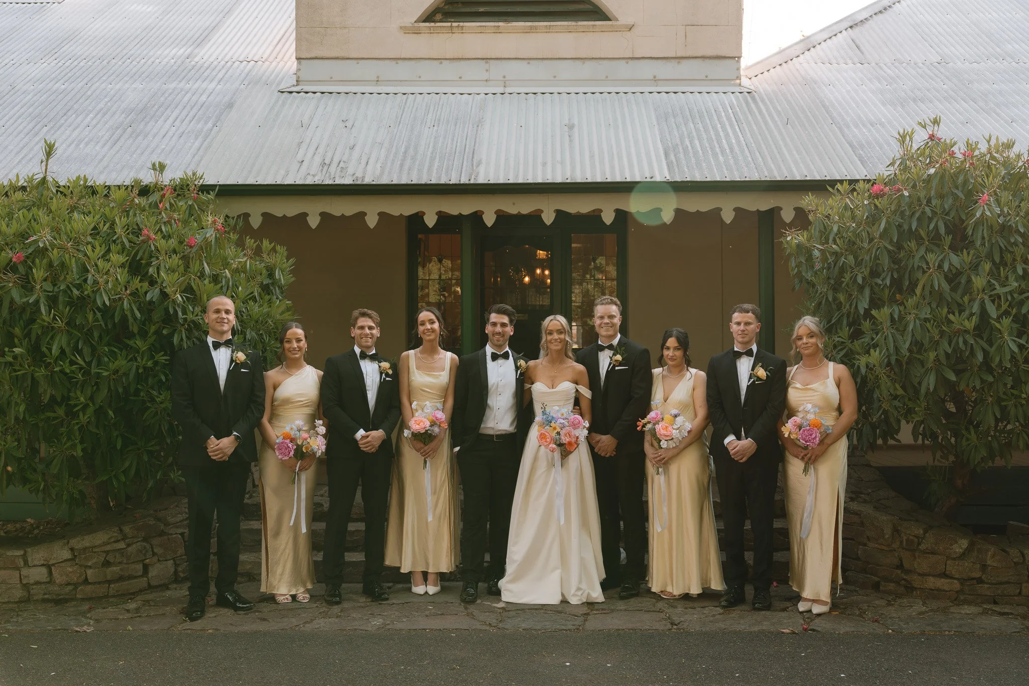 Group of ten people dressed in formal wedding attire standing outside in front of a building with trees on each side. The bride and groom are in the center, surrounded by bridesmaids and groomsmen.