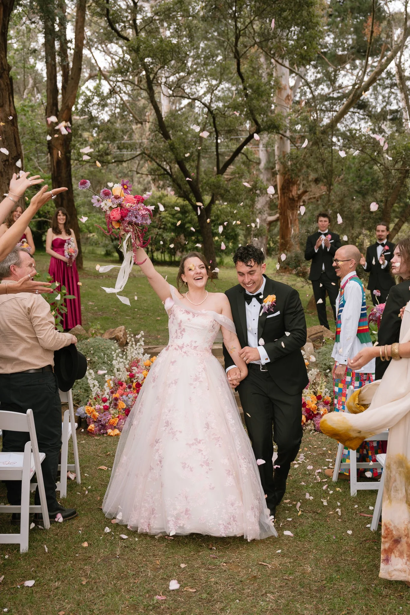 A bride and groom celebrating their wedding outdoors, walking hand in hand, with guests throwing flower petals around them.