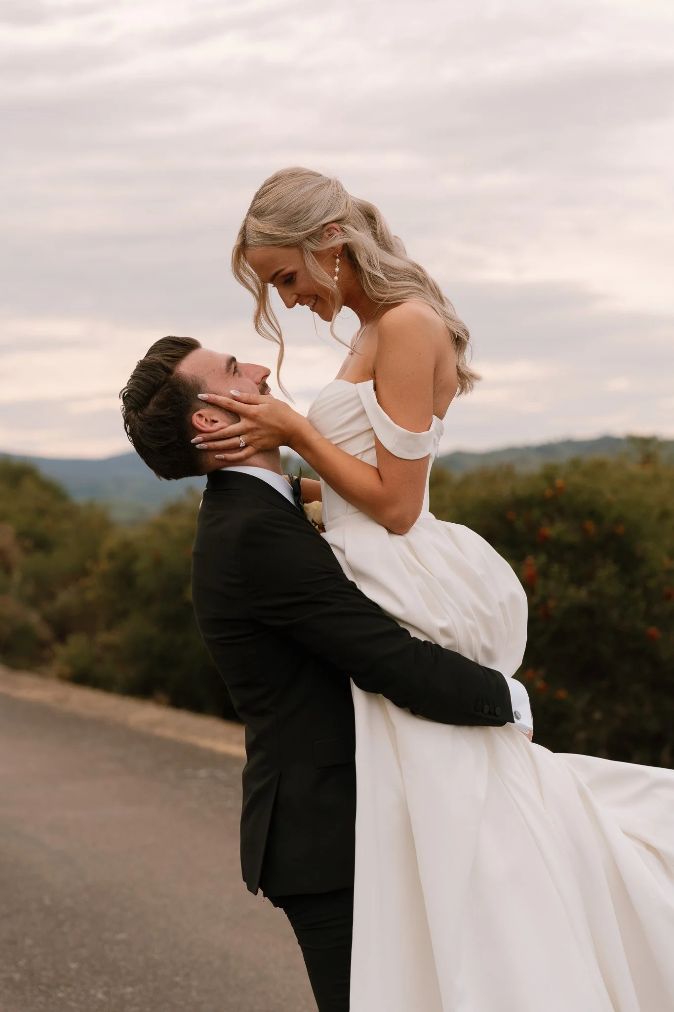 A bride and groom celebrate outdoors, the groom holding the bride up as she smiles down at him, with a scenic landscape and cloudy sky in the background.