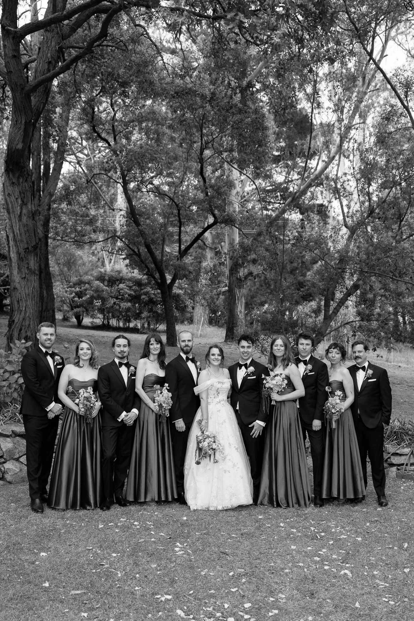 Black and white photo of a wedding party outdoors. The bride and groom are in the center, with the bride holding a bouquet and wearing a white wedding dress. The groom wears a tuxedo. They are surrounded by six bridesmaids and groomsmen dressed in fo