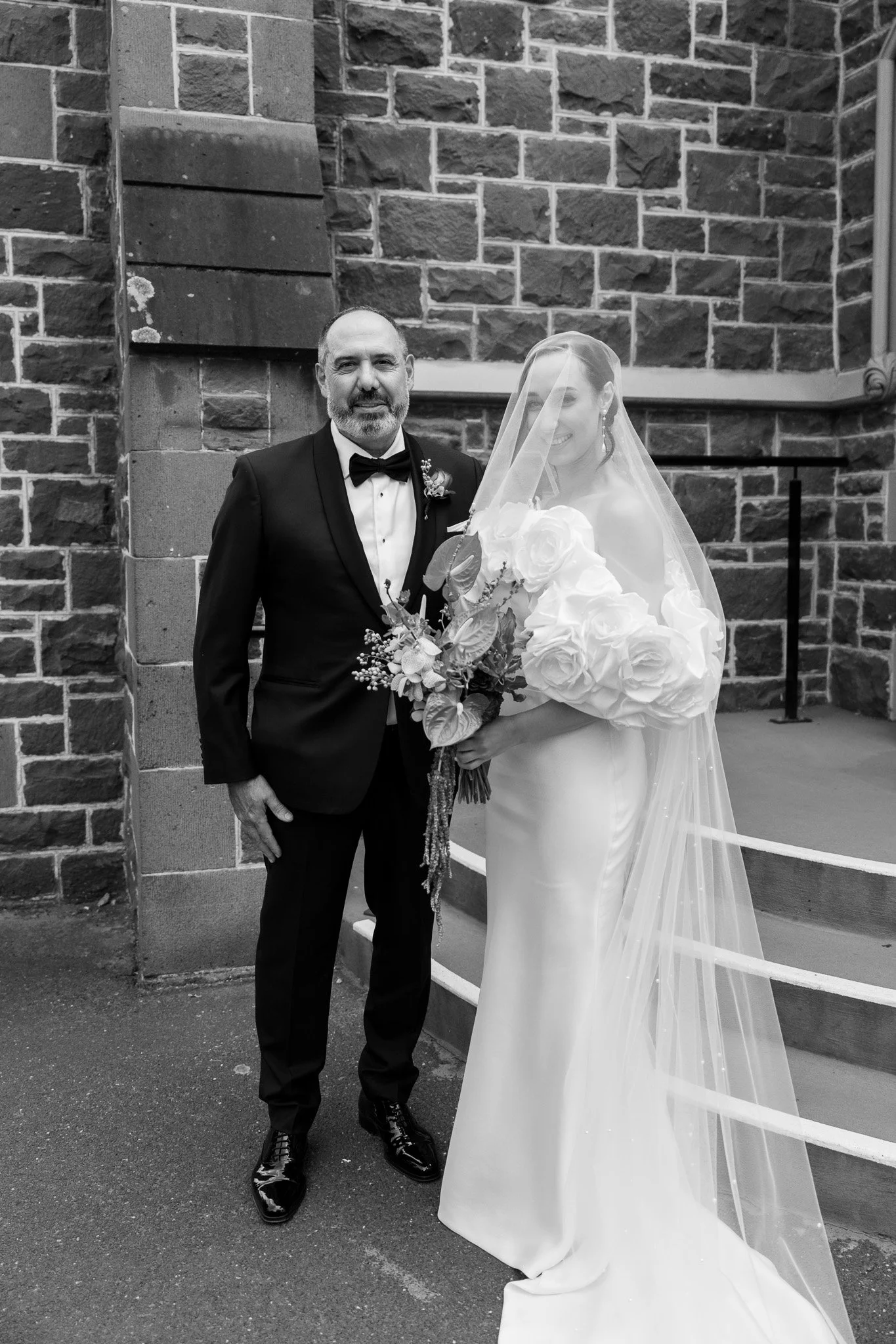 Black and white photo of a bride and a man in a tuxedo, standing outdoors in front of a brick building. The bride is holding a bouquet of roses and wrapped in a long veil.