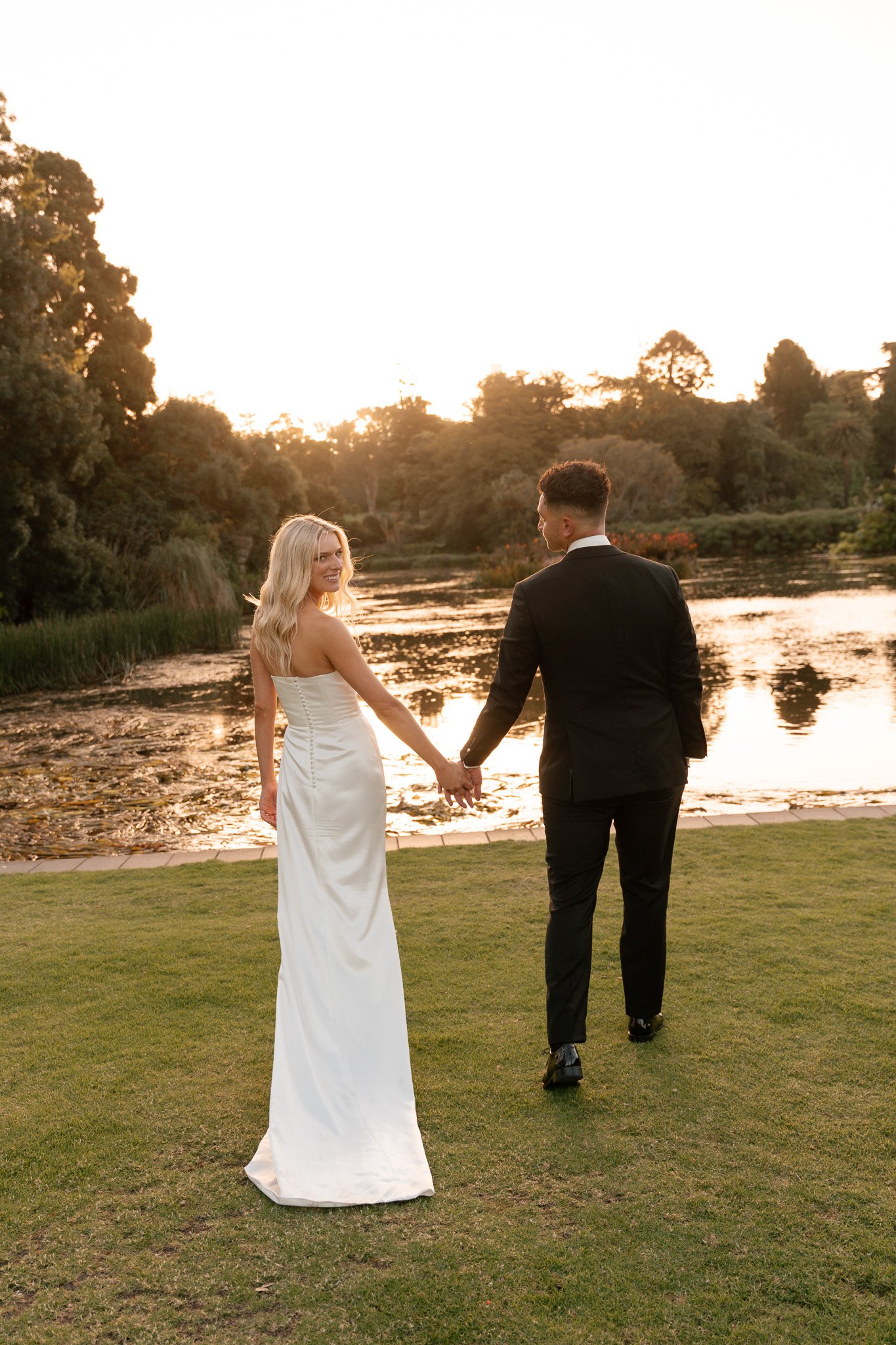 A bride and groom holding hands, walking by a lake at sunset, with trees in the background.