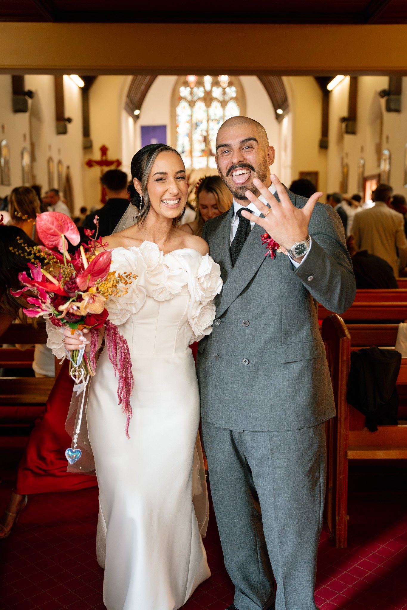 Bride in white wedding gown holding a bouquet, smiling, standing next to groom in gray suit, showing off wedding ring, inside a church with stained glass window in background.