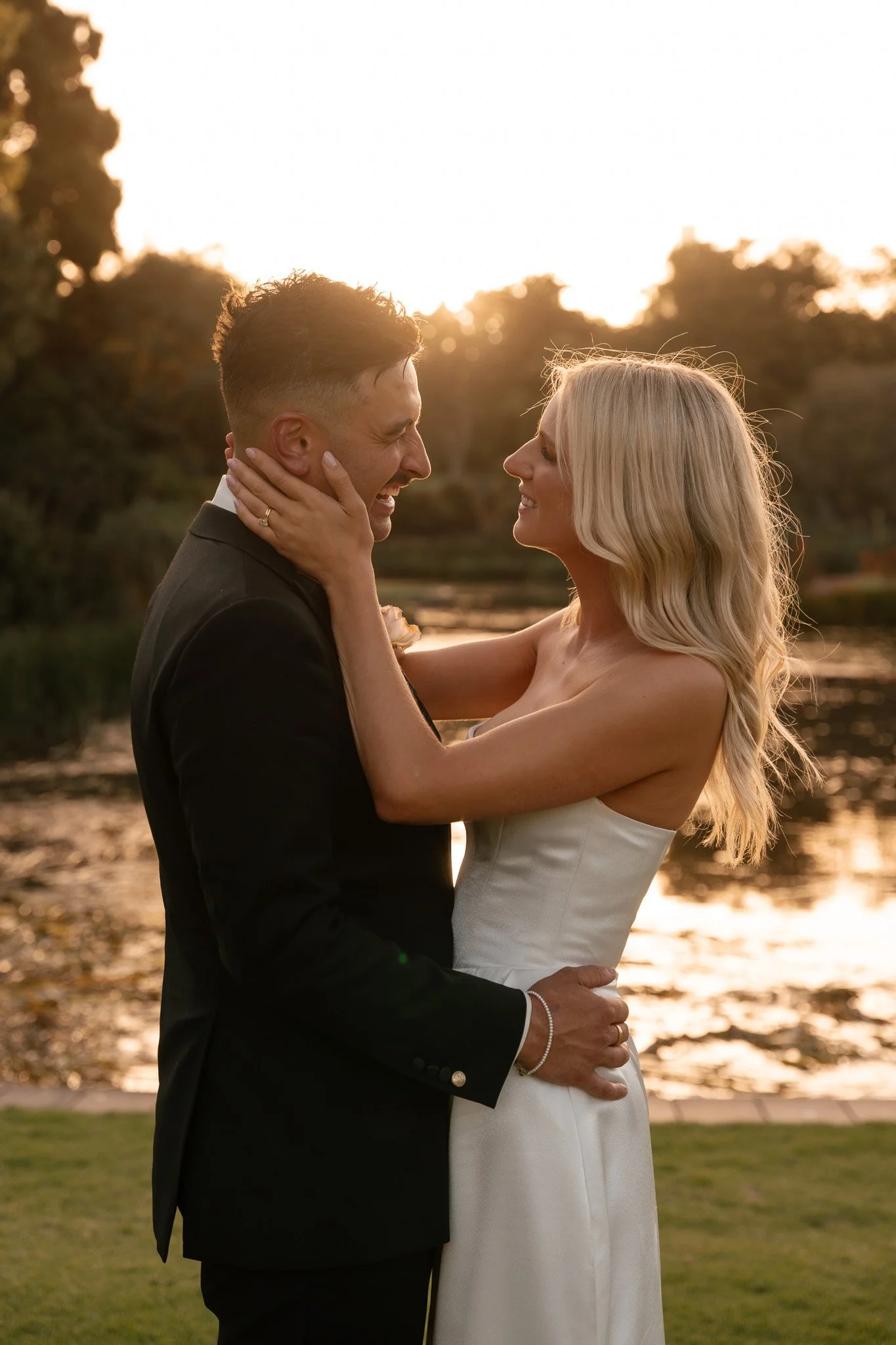 A newlywed couple is embracing outdoors at sunset, with the bride in a strapless wedding dress and the groom in a black suit, near a body of water surrounded by trees.