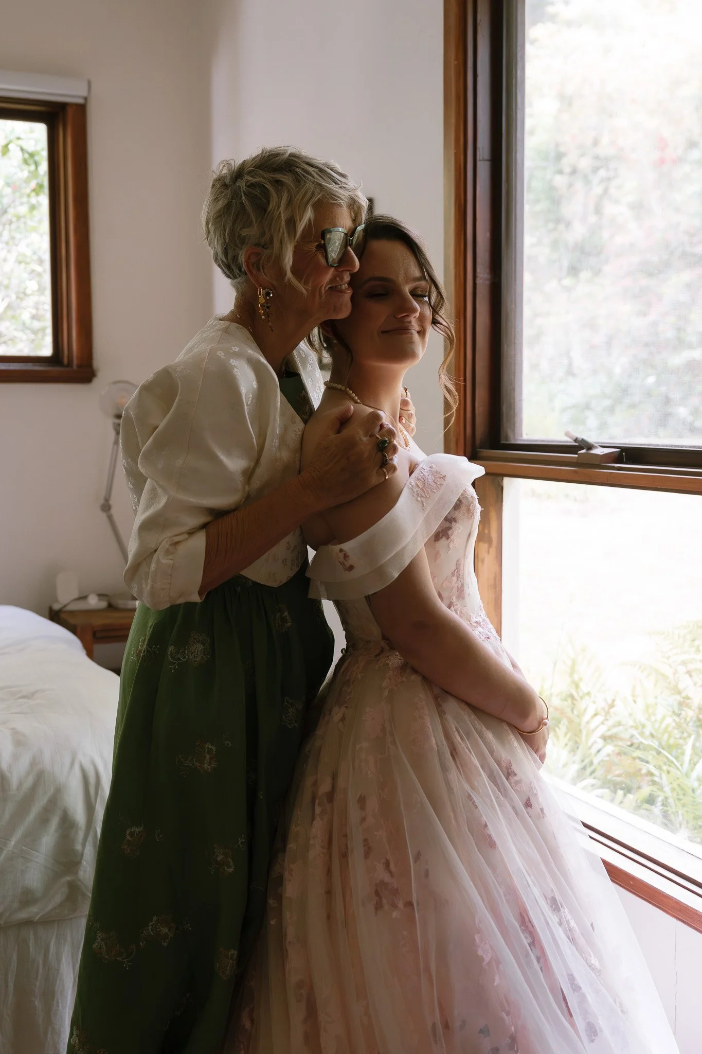 A woman and an elderly woman embracing by a window, with the younger woman wearing a pink wedding dress with floral details and the older woman wearing glasses and a green skirt, in a softly lit room.
