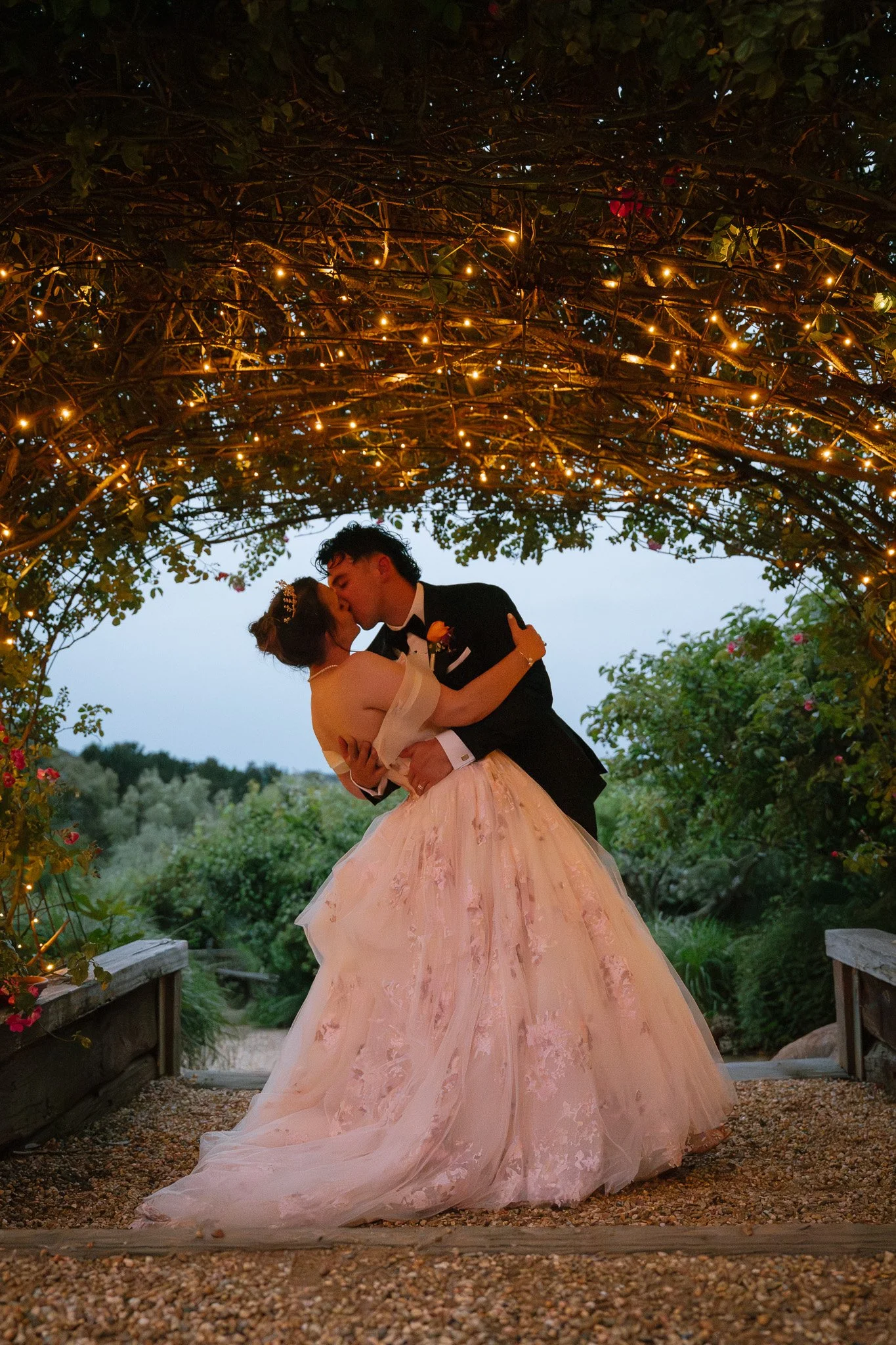 A bride and groom sharing a kiss under a lit, arching trellis at sunset.
