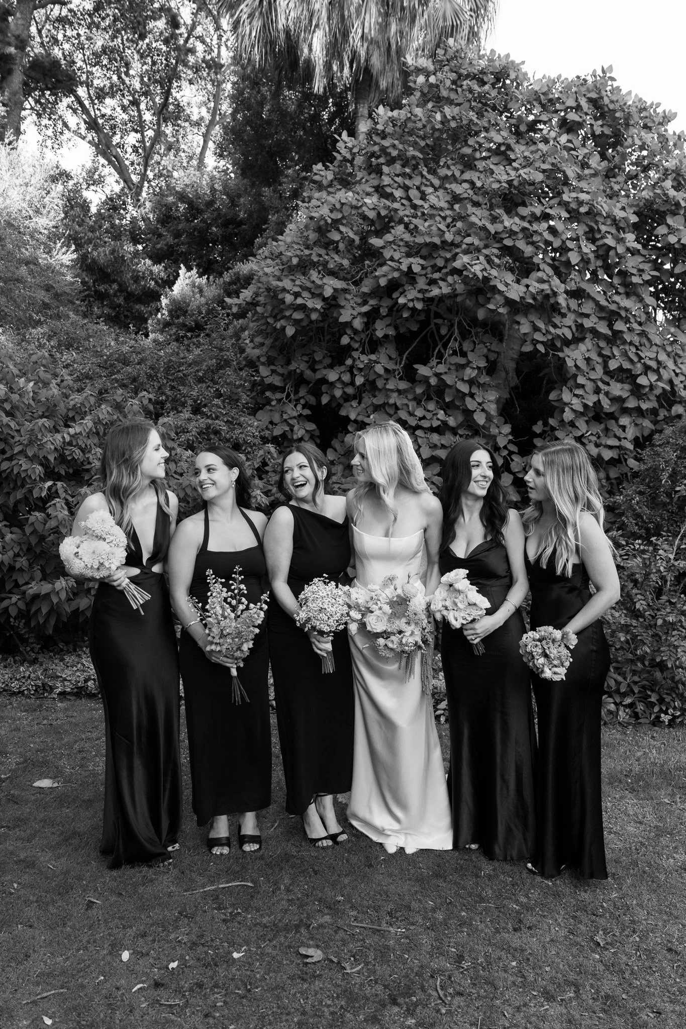 Black-and-white photo of six women dressed in formal gowns standing together outside near trees, smiling and holding bouquets of flowers.