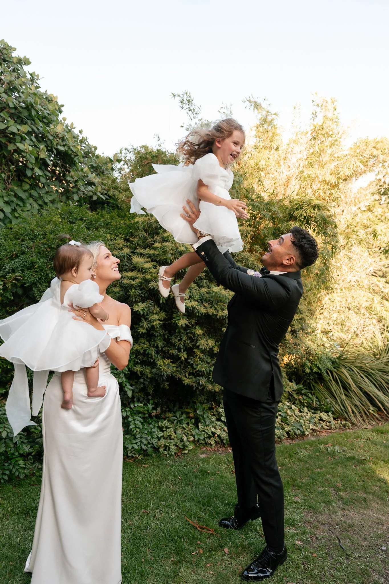 A man in a black tuxedo lifts a young girl in a white dress high into the air while a woman in a white dress with another young girl in a white dress look on joyfully, all outdoors with greenery and trees in the background.