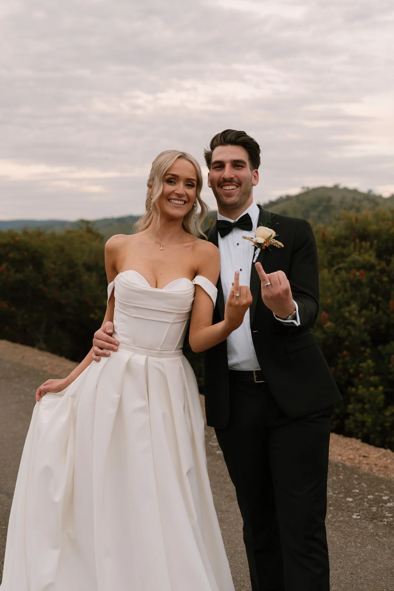 A happy couple on their wedding day standing outdoors. The bride is wearing a white off-shoulder wedding gown, and the groom is dressed in a black tuxedo with a bow tie. Both are smiling and making gestures with their hands.