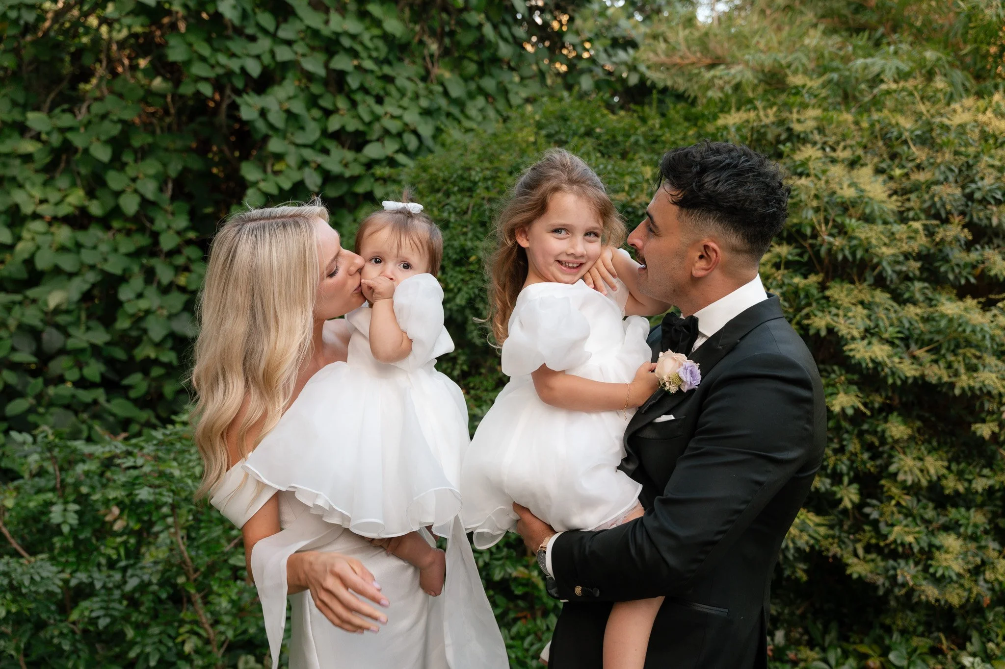 A family of four dressed in formal wedding attire outdoors, with a lush green background. The mother is kissing one young girl, while the father holds the other girl, who is smiling.