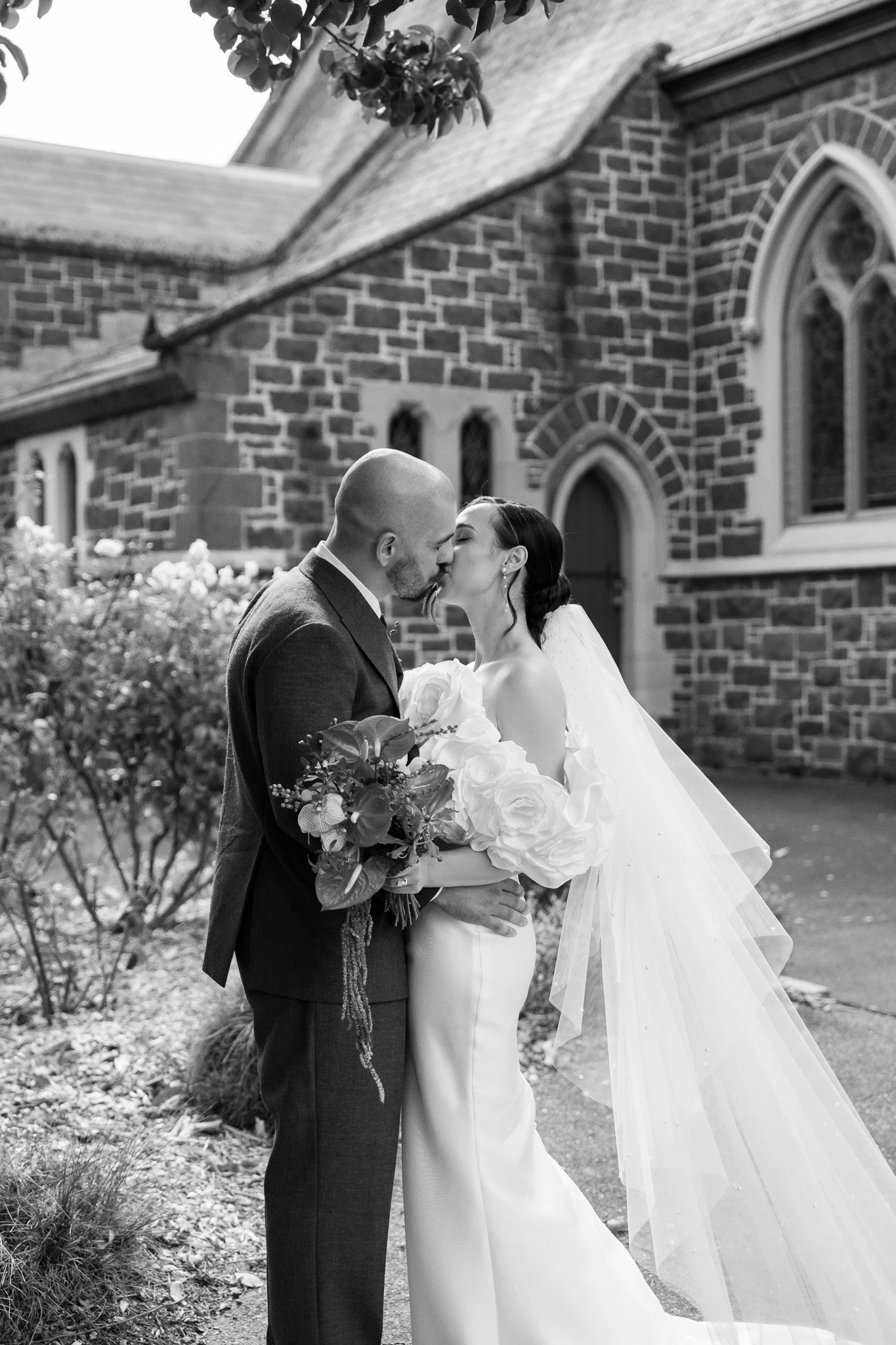 Black and white photo of a bride and groom kissing outside a church, with the bride holding a bouquet of flowers and wearing a veil, and the groom dressed in a suit.