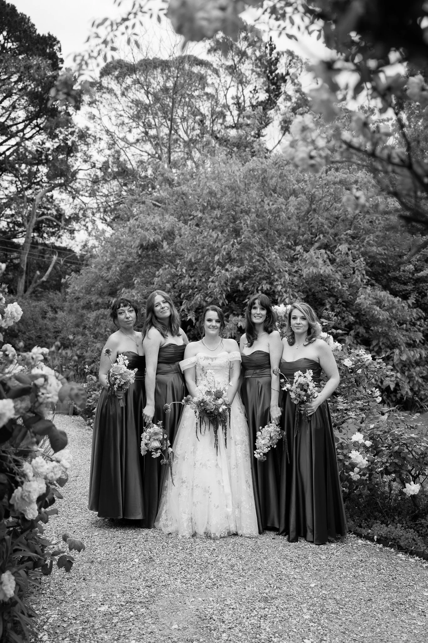 Black and white photo of five women in formal dresses standing on a gravel path in a garden with dense trees and bushes.