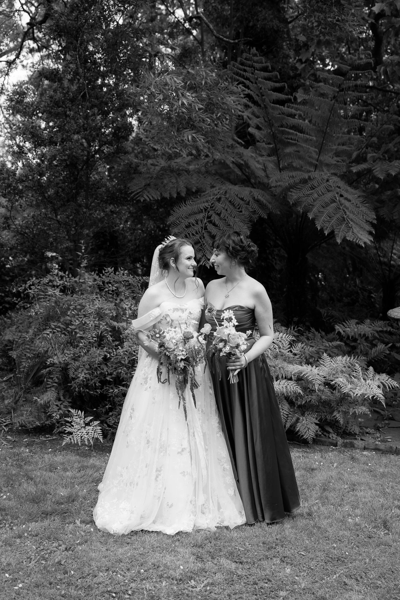 Black and white photo of two women in wedding attire standing together outdoors amidst trees and ferns, holding bouquets and smiling at each other.