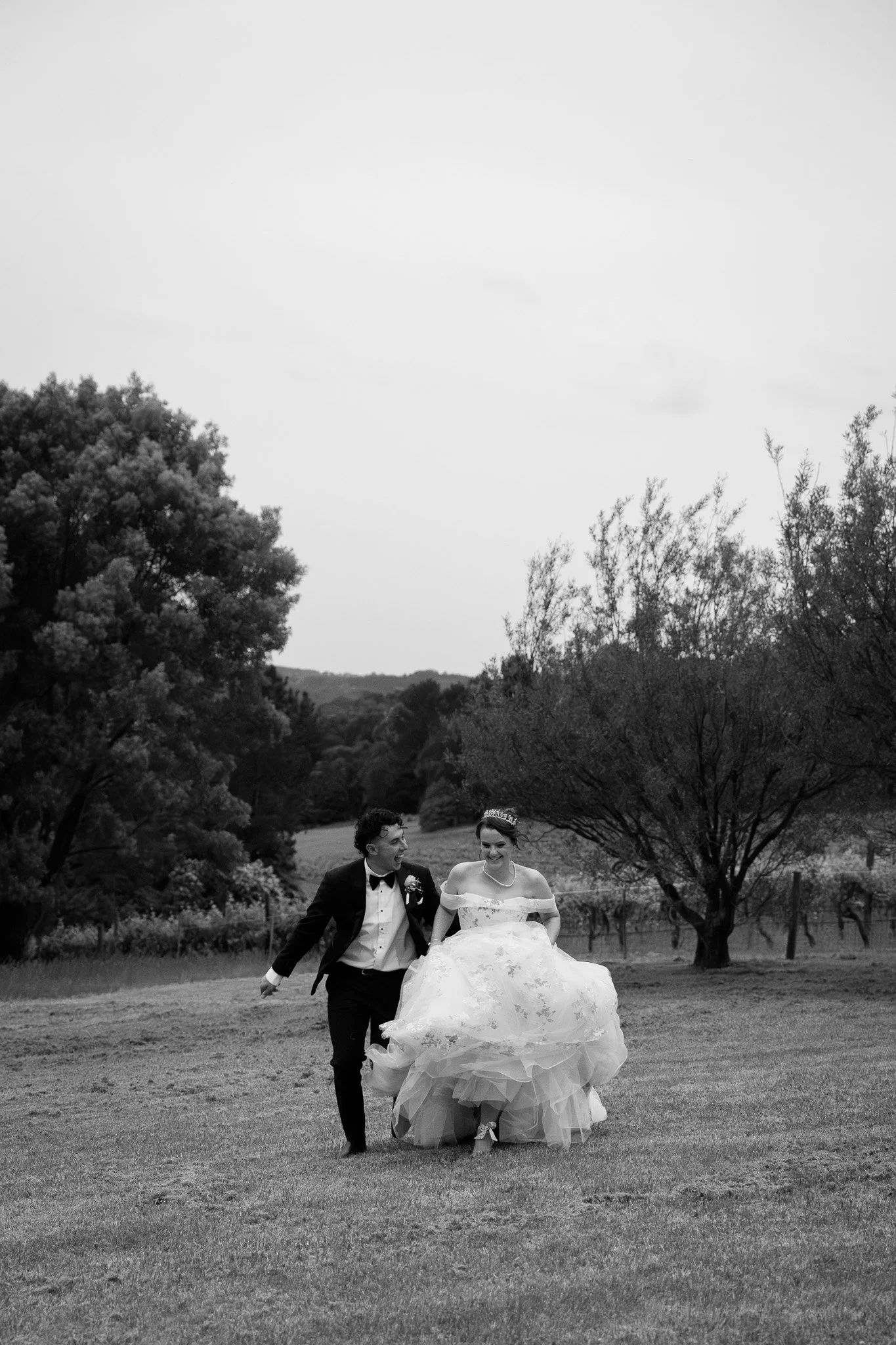 A black and white photo of a bride and groom walking in a field, smiling and holding hands, with trees in the background.