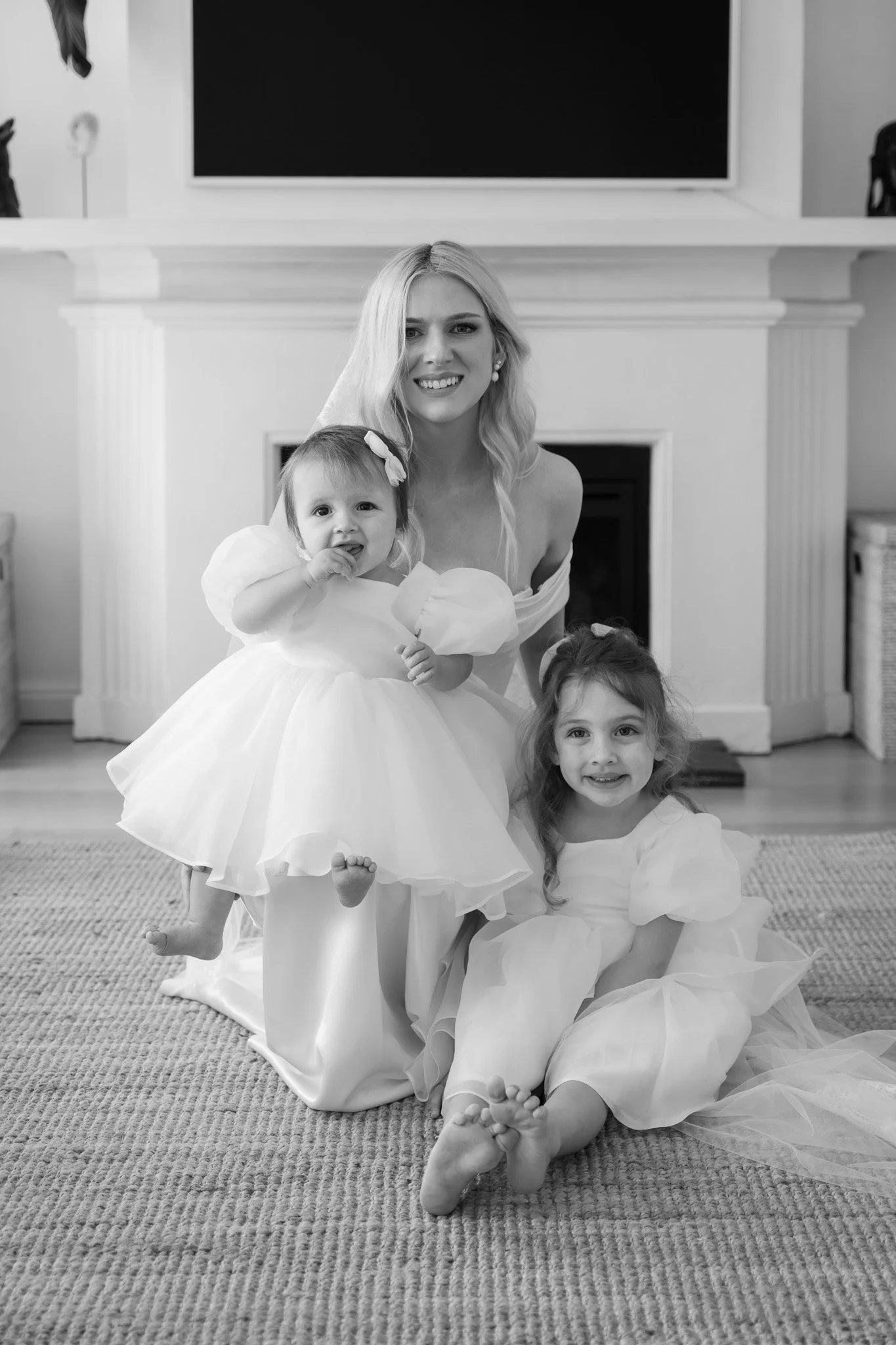 Black and white photo of a woman with two young girls dressed in white dresses, sitting on a carpeted floor in front of a fireplace, smiling at the camera.