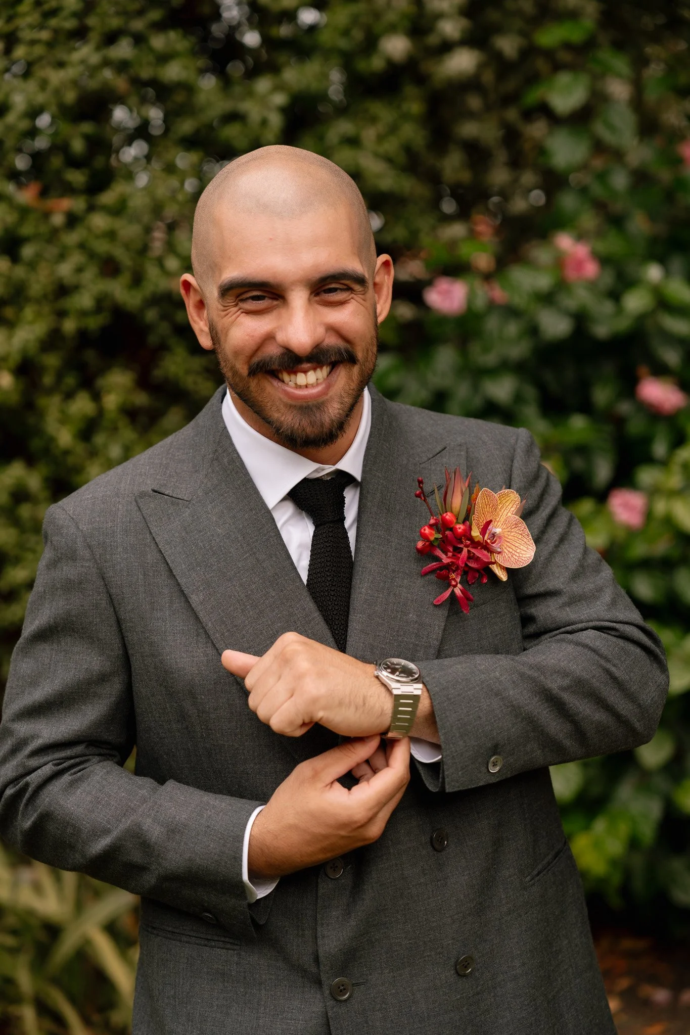 A man in a gray suit and black tie smiling outdoors, wearing a wristwatch and a boutonniere with pink and red flowers on his left lapel.