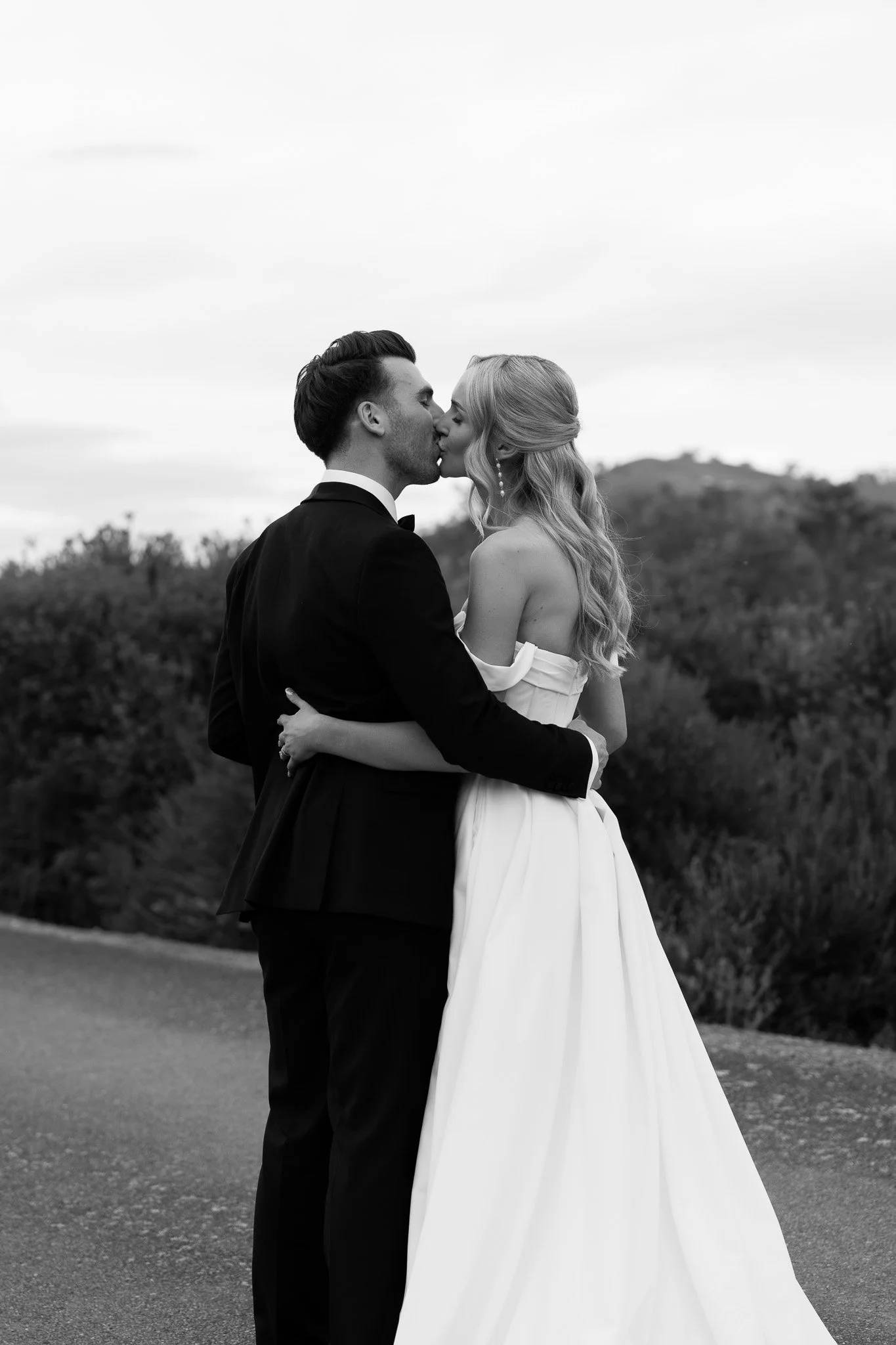 A black-and-white photo of a bride and groom kissing outdoors, with hills and trees in the background.