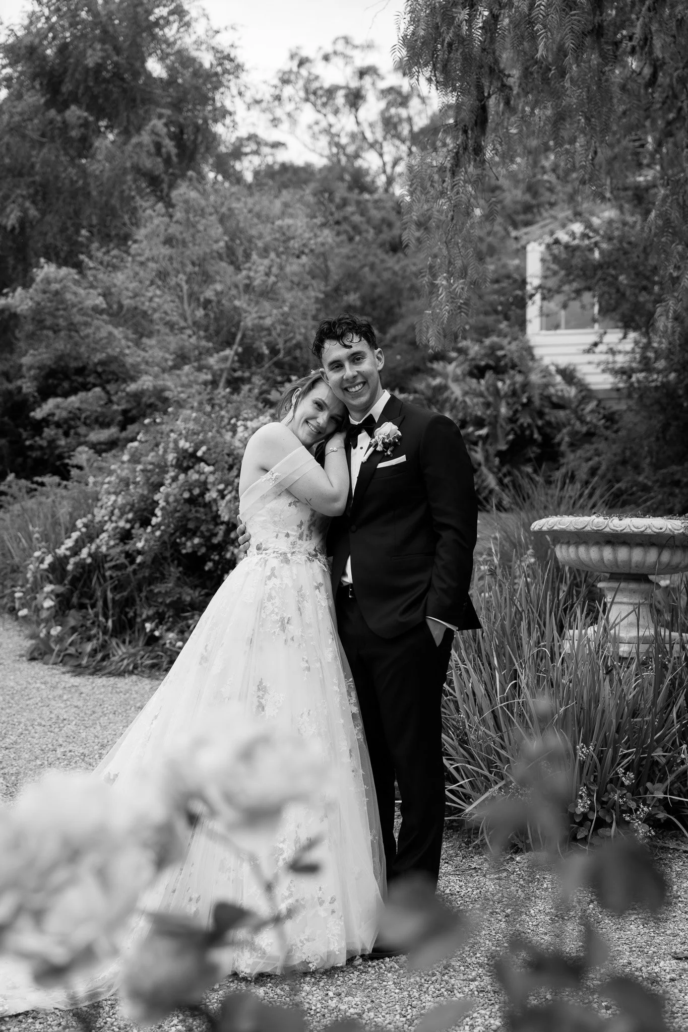 A black and white photo of a happy couple, the bride in a wedding gown and the groom in a tuxedo, embracing each other outdoors among trees and plants.