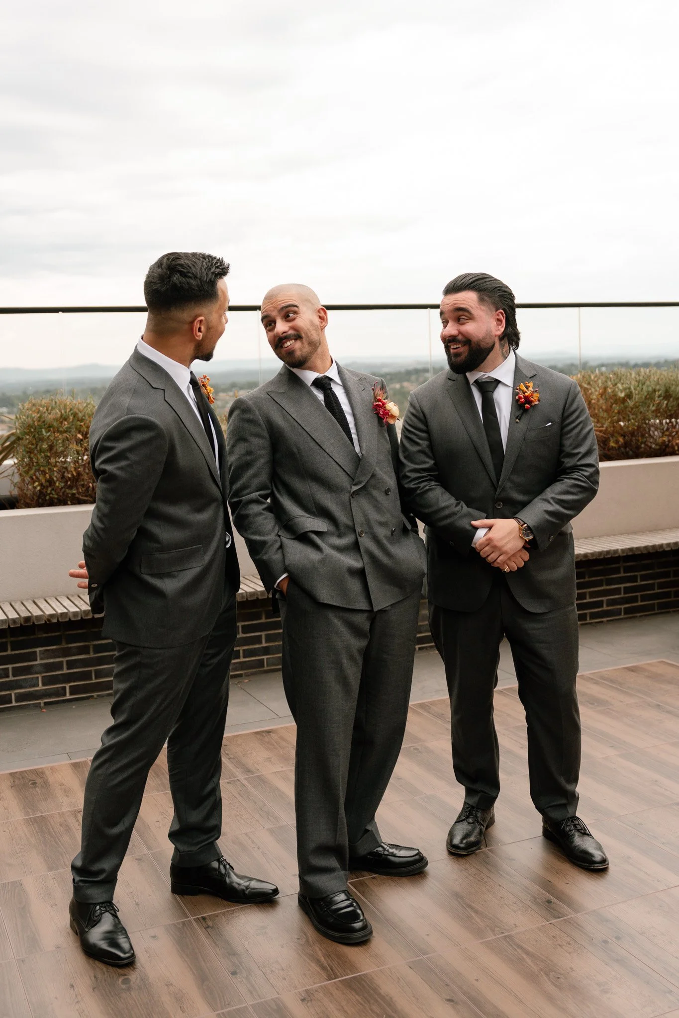 Three men in gray suits having a conversation on a rooftop terrace with a scenic view in the background.